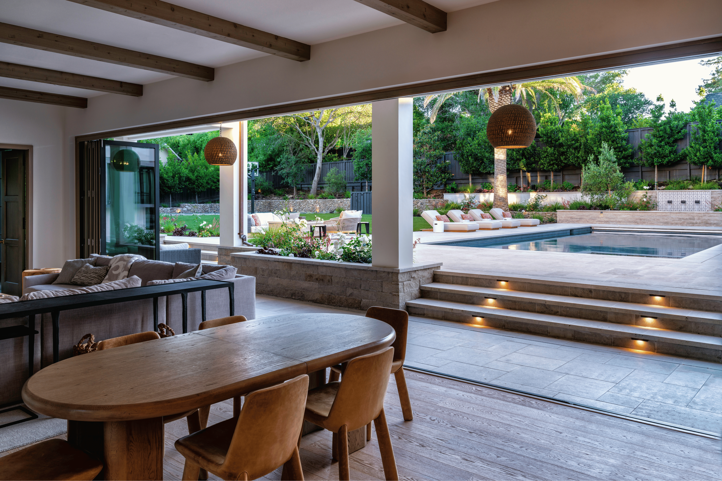 View of a modern open-concept living and dining area opening to a backyard with a pool, lounge chairs, greenery, and trees.