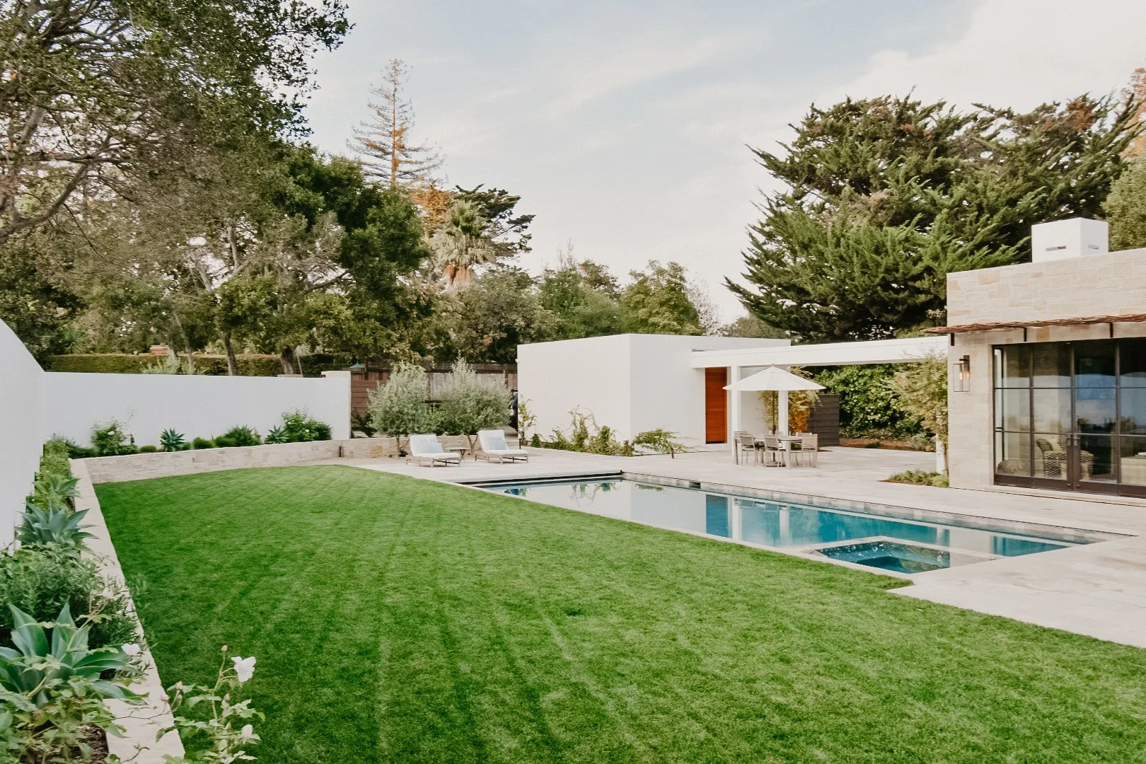 Modern backyard with a swimming pool, grassy lawn, and outdoor dining area under an umbrella, surrounded by trees and white walls.
