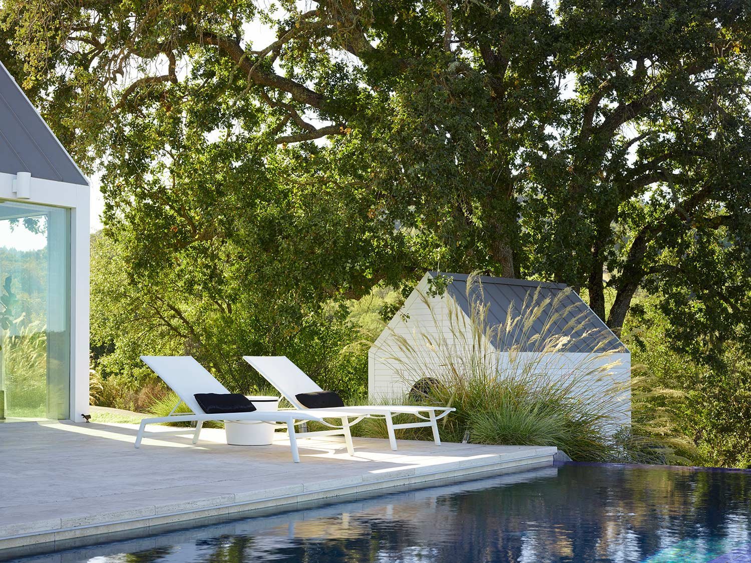 A modern outdoor pool area with two white lounge chairs and black cushions, a small white table, a white dog house, and lush green trees in the background.