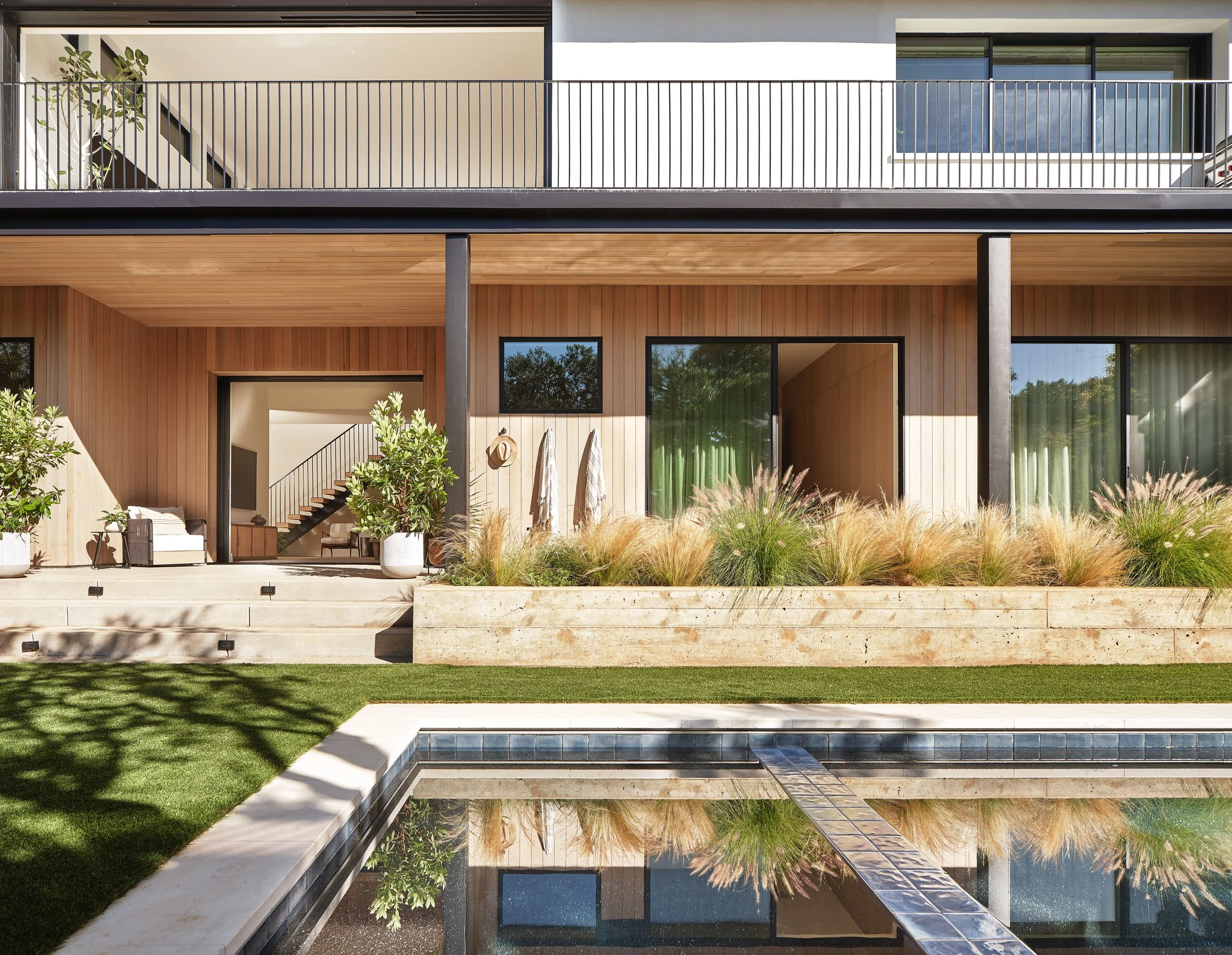 Modern house with a wooden facade, large glass sliding doors, front patio, lush landscaping, and a swimming pool in the foreground.