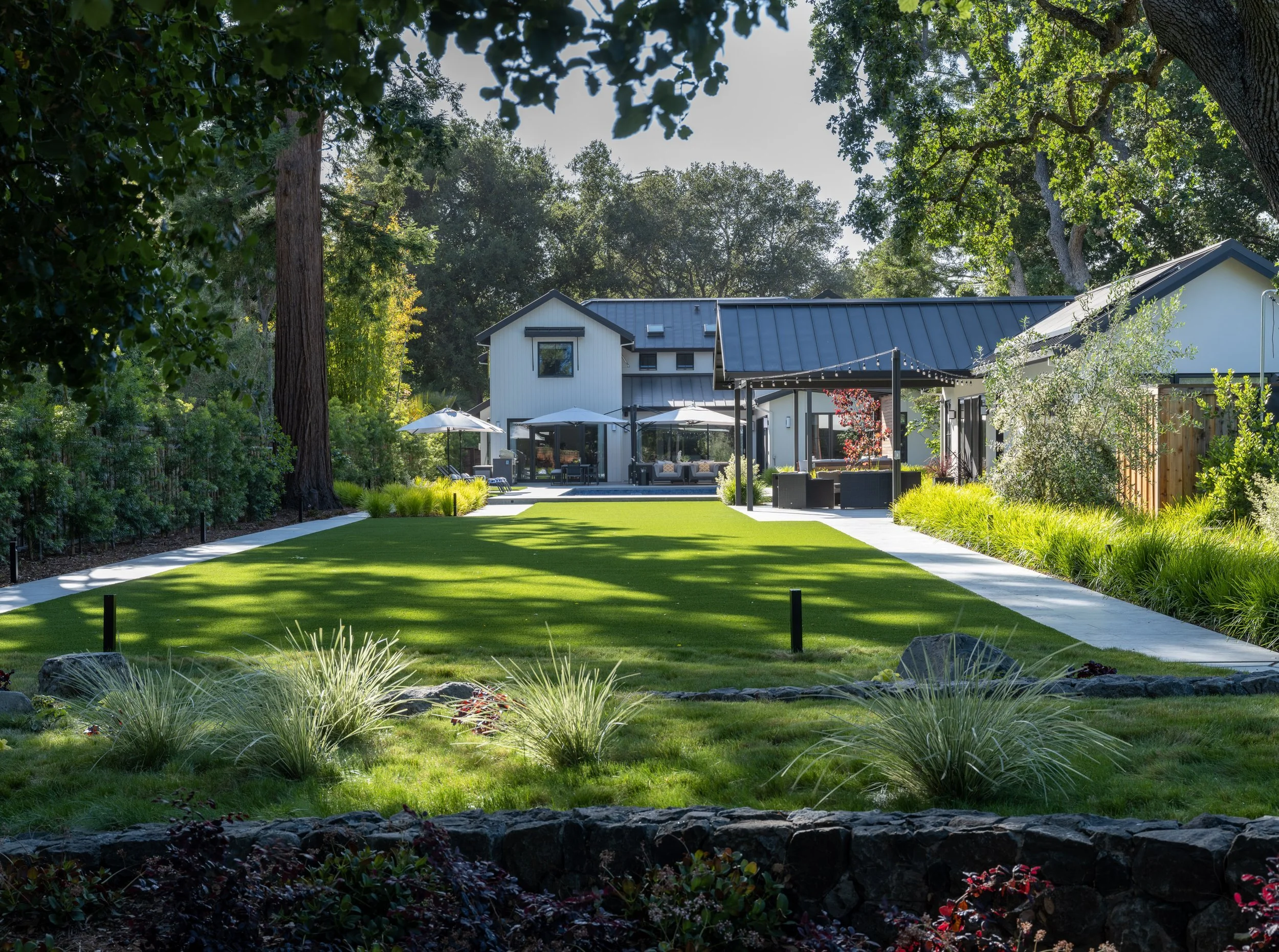 A well-maintained backyard with green grass, landscaping, trees, and a modern house in the background, featuring outdoor seating with umbrellas and a patio.