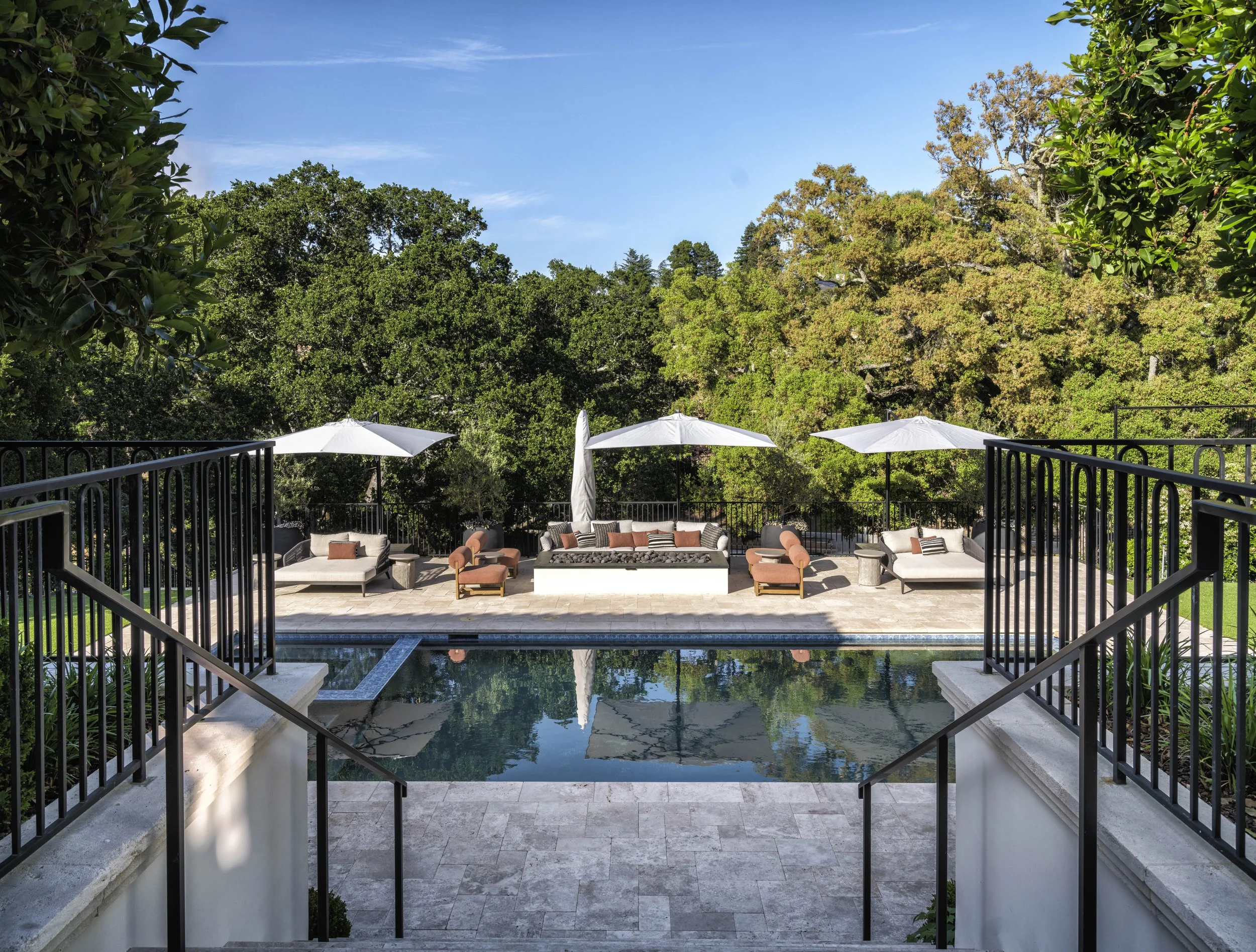 Outdoor pool area with lounge chairs, umbrellas, and seating on a tiled deck, surrounded by green trees under a blue sky.