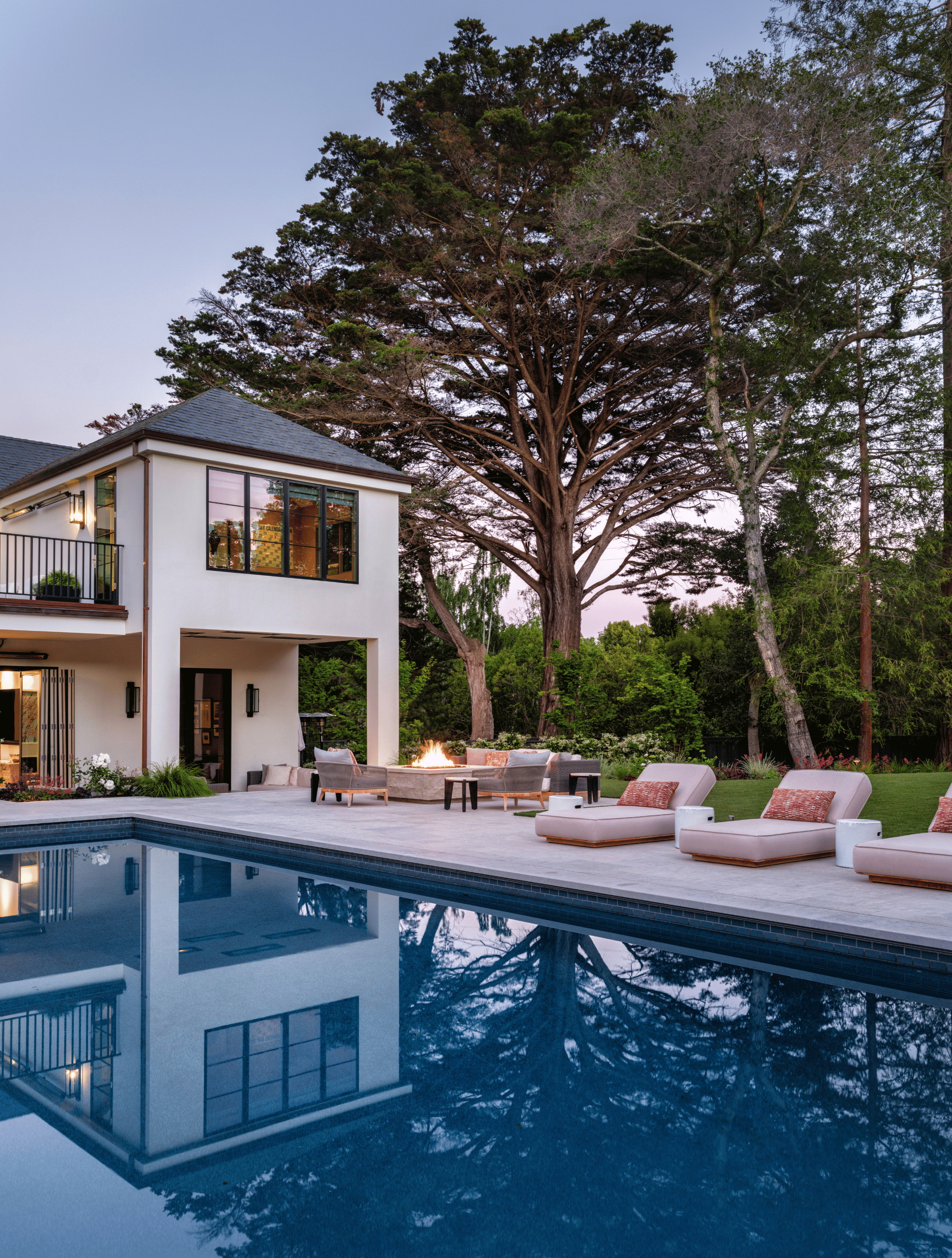 Modern backyard with swimming pool, outdoor seating area with couches and fire pit, large tree, and a white house with large windows at dusk.