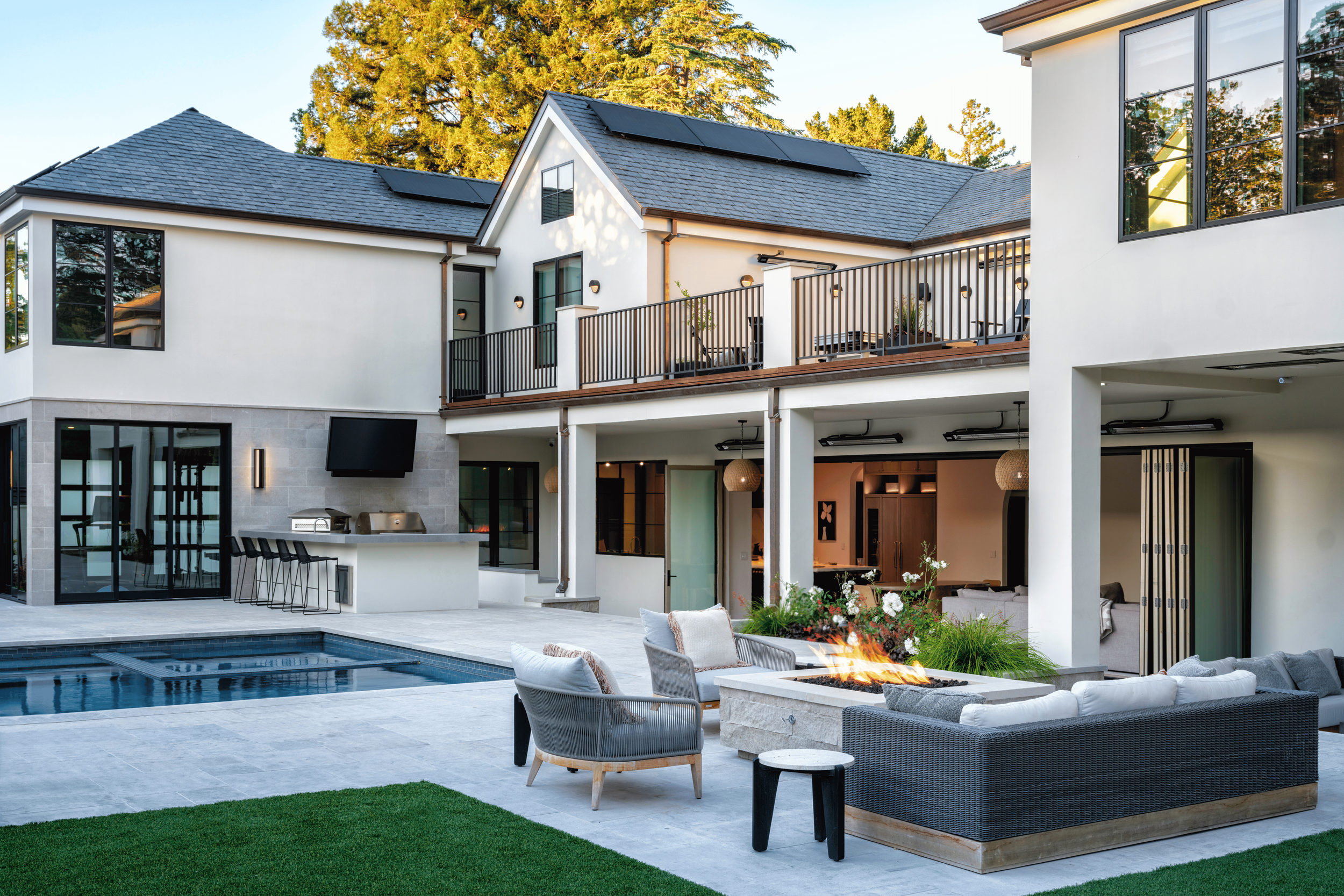 A modern backyard with a swimming pool, outdoor seating with fire pit, and a two-story house with large windows and an upper balcony.