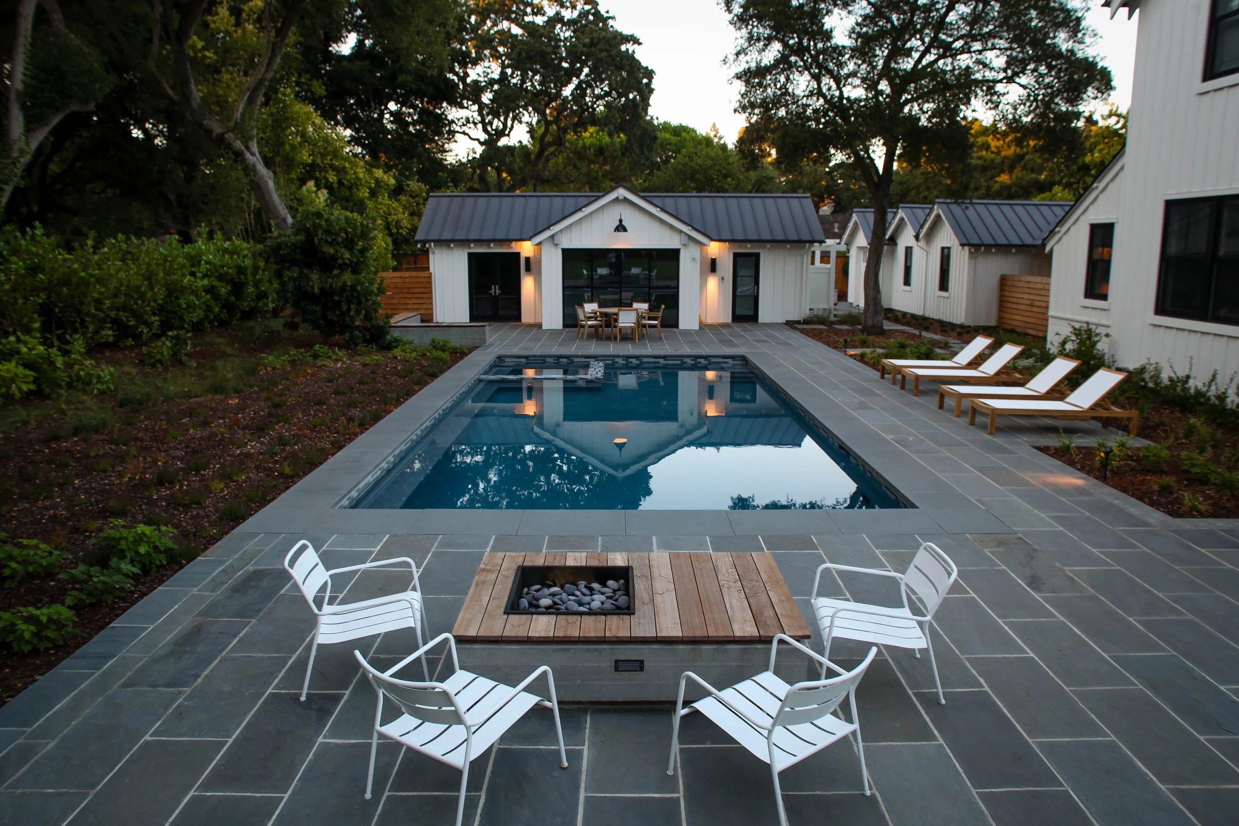 Modern backyard with a rectangular swimming pool, surrounded by grey stone pavers. There are six white chairs around a fire pit in the foreground and four white lounge chairs along the right side. At the back, there is a white pool house with a black