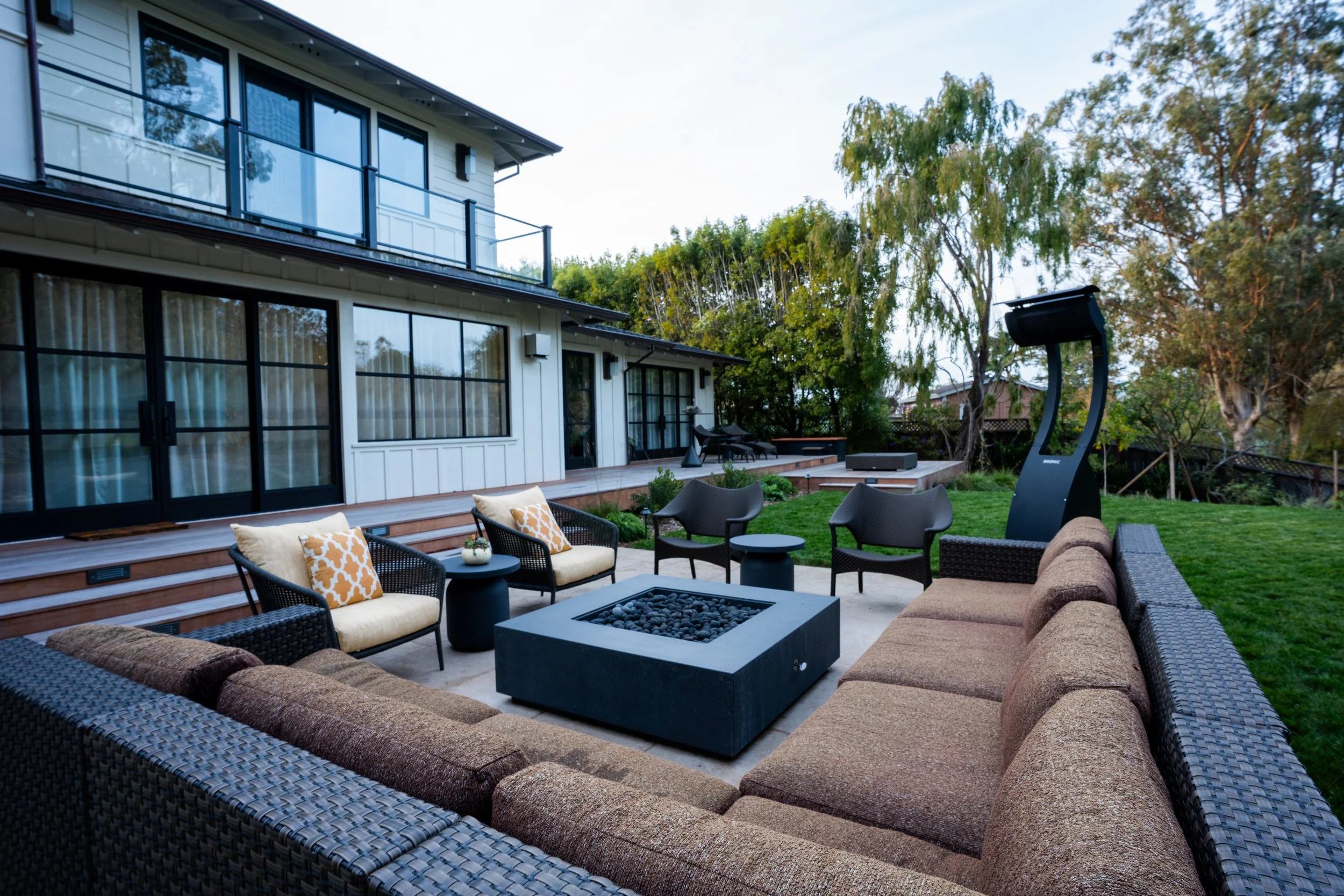 Outdoor patio with a large sectional sofa, several armchairs, a fire pit, and a tall outdoor heater in a backyard with a grassy lawn, trees, and a modern two-story house with large windows in the background.