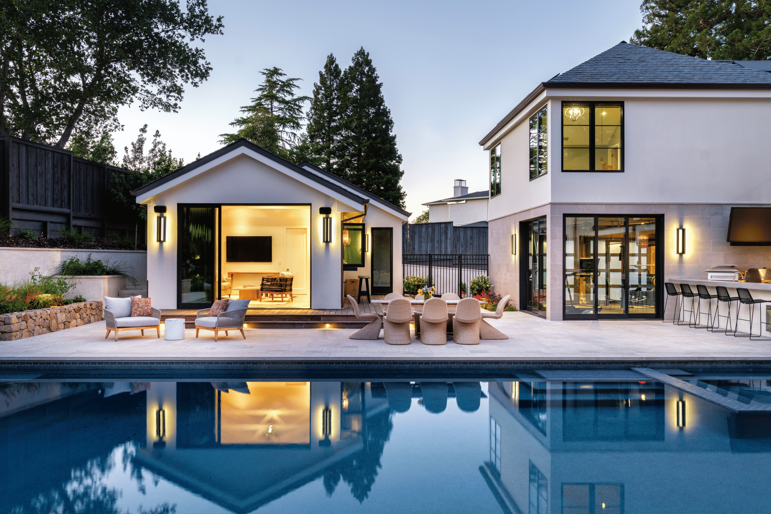 Modern backyard with pool, outdoor seating, and a small house with large glass doors and windows, illuminated by outdoor lights at dusk.