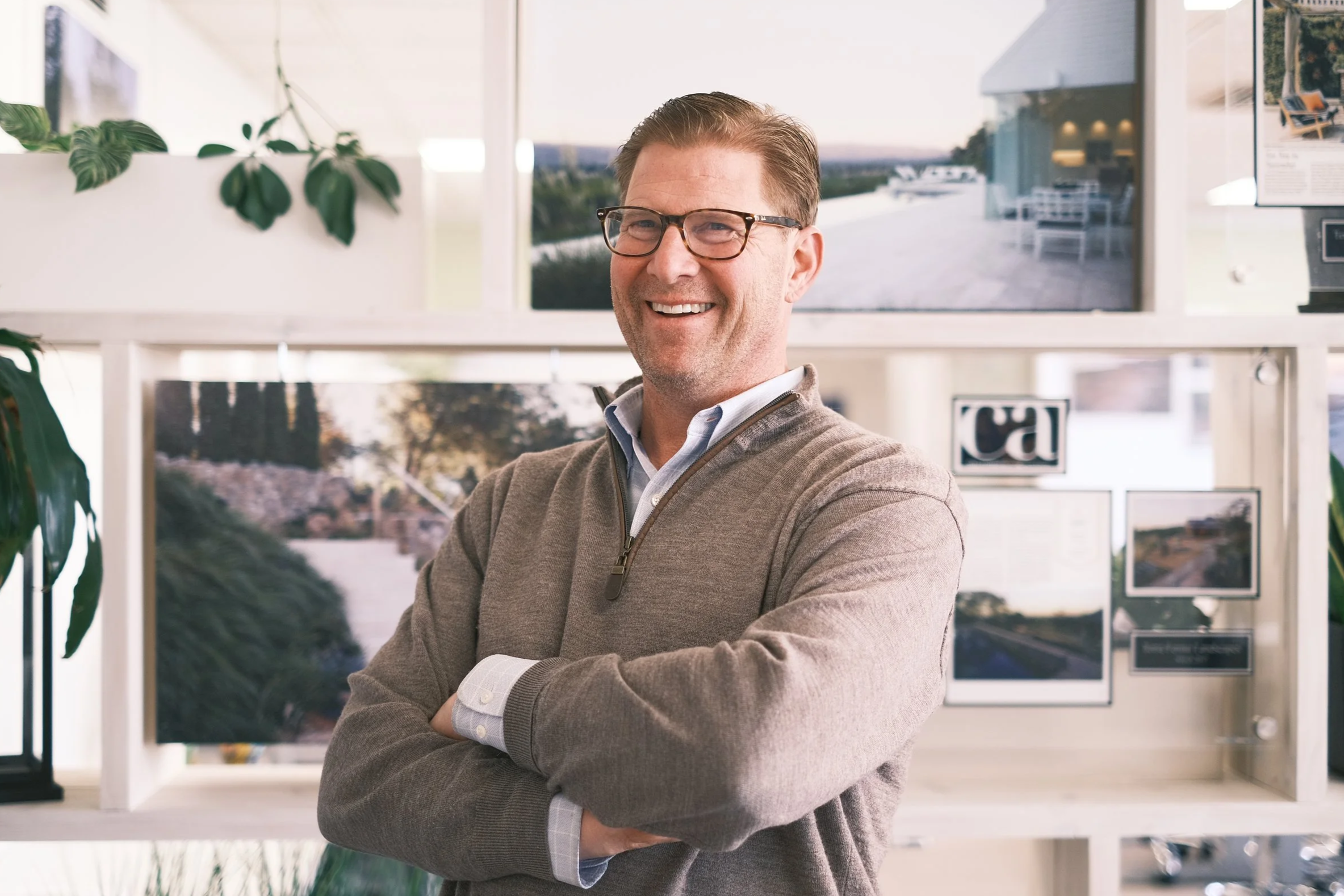 A man smiling in an indoor office setting with plants and monitors in the background.