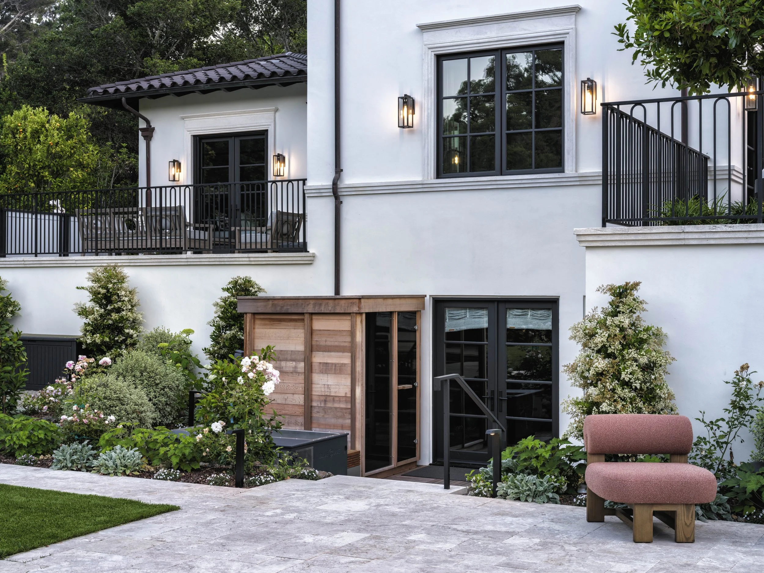 Exterior view of a modern white house with black-framed windows and doors, lit wall-mounted lights, a small wooden shed, a patio with a pink cushioned chair, and lush green landscaping with flowering bushes.
