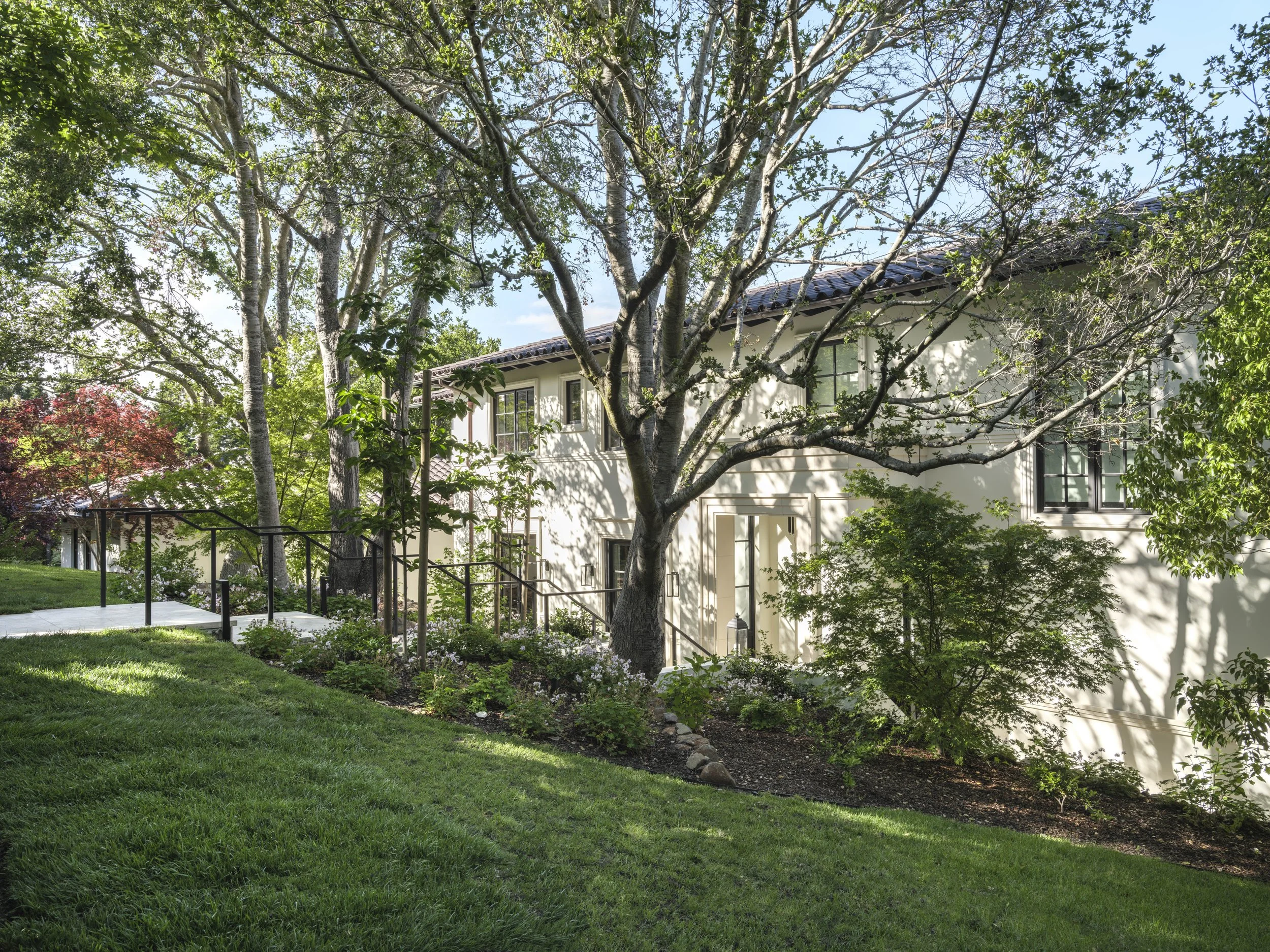 A white two-story house with multiple windows, surrounded by lush green trees and shrubs. A landscaped garden with a grass lawn and a concrete walkway with black metal railings is visible in the foreground, casting shadows on the house.