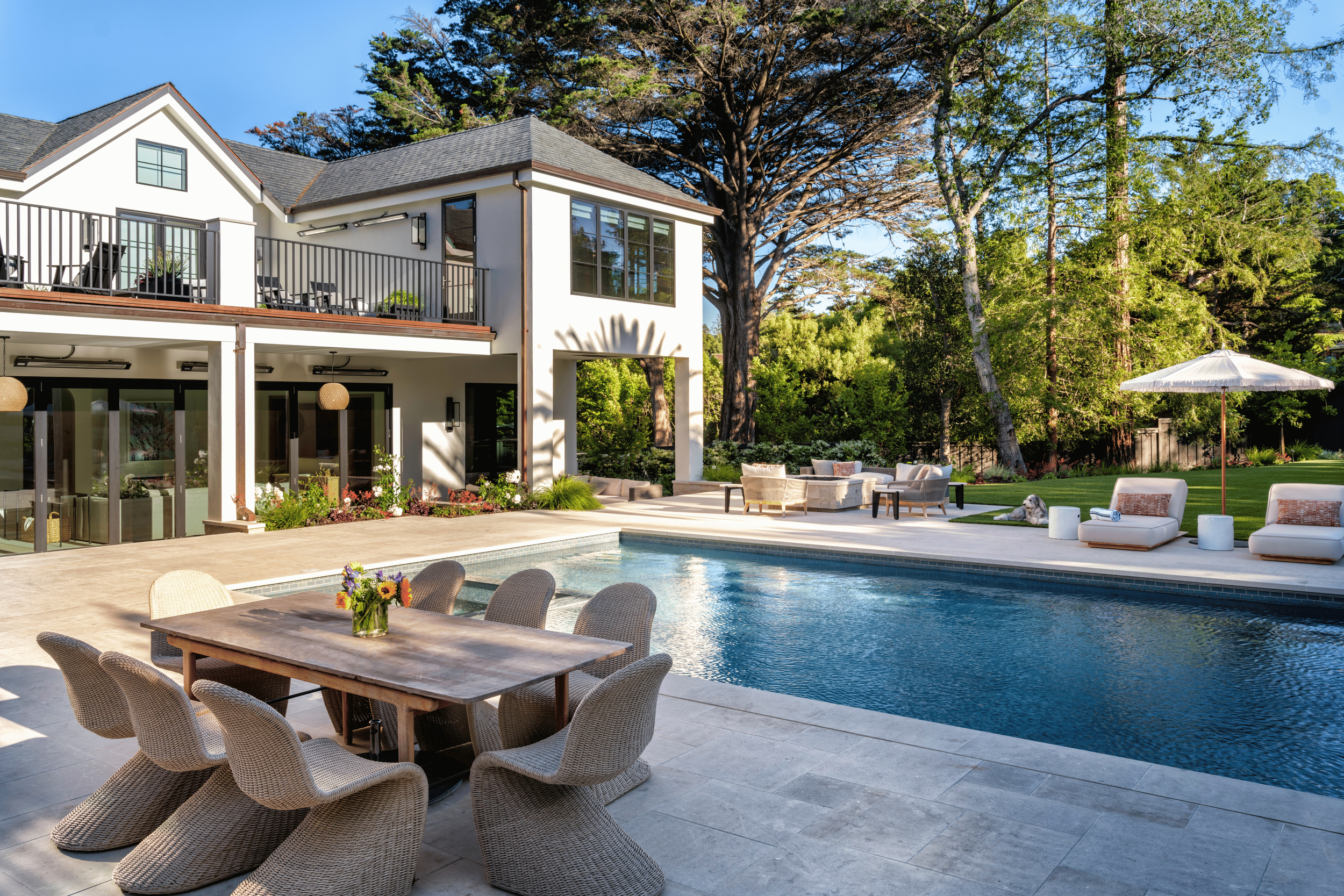 Porch area with outdoor furniture, swimming pool, garden with lush green trees, sun umbrellas, and a two-story house with large windows and a balcony.