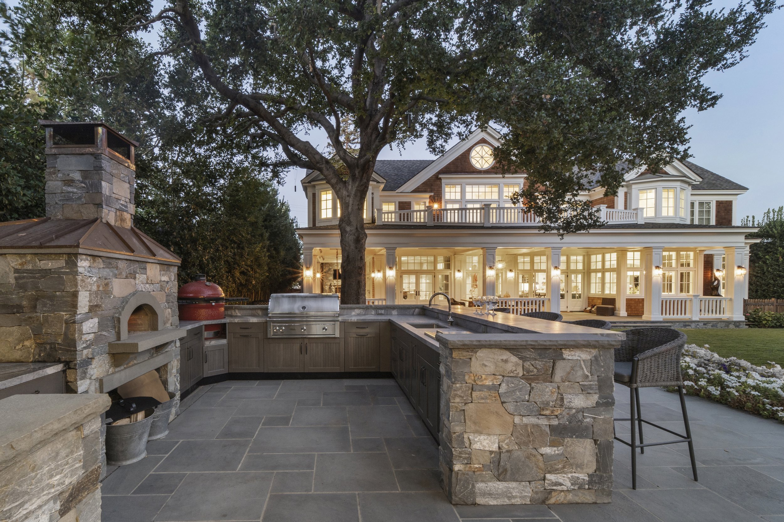 Outdoor kitchen in a backyard with stone countertops, built-in grill, and a fireplace, overlooking a large lit house with multiple windows and a balcony at dusk.