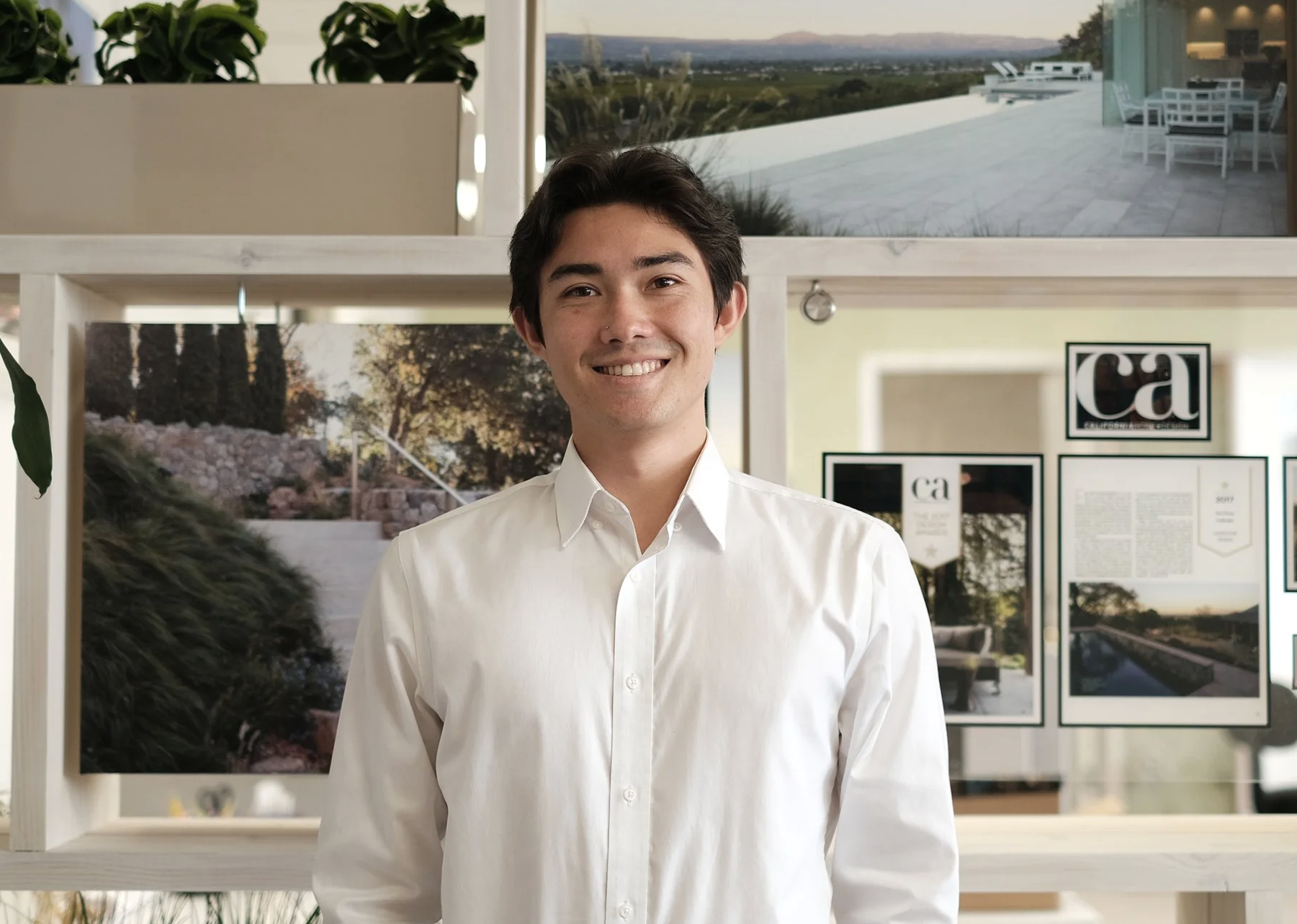 A young man with dark hair, wearing a white button-up shirt, standing indoors with a smile.