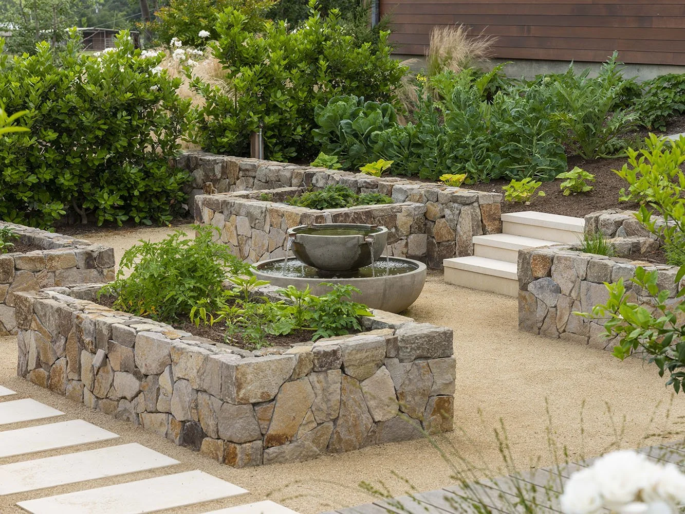 A landscaped garden with stone planters filled with green plants, a small tiered water fountain, white steps leading up a slope, and lush shrubs and garden beds in the background.