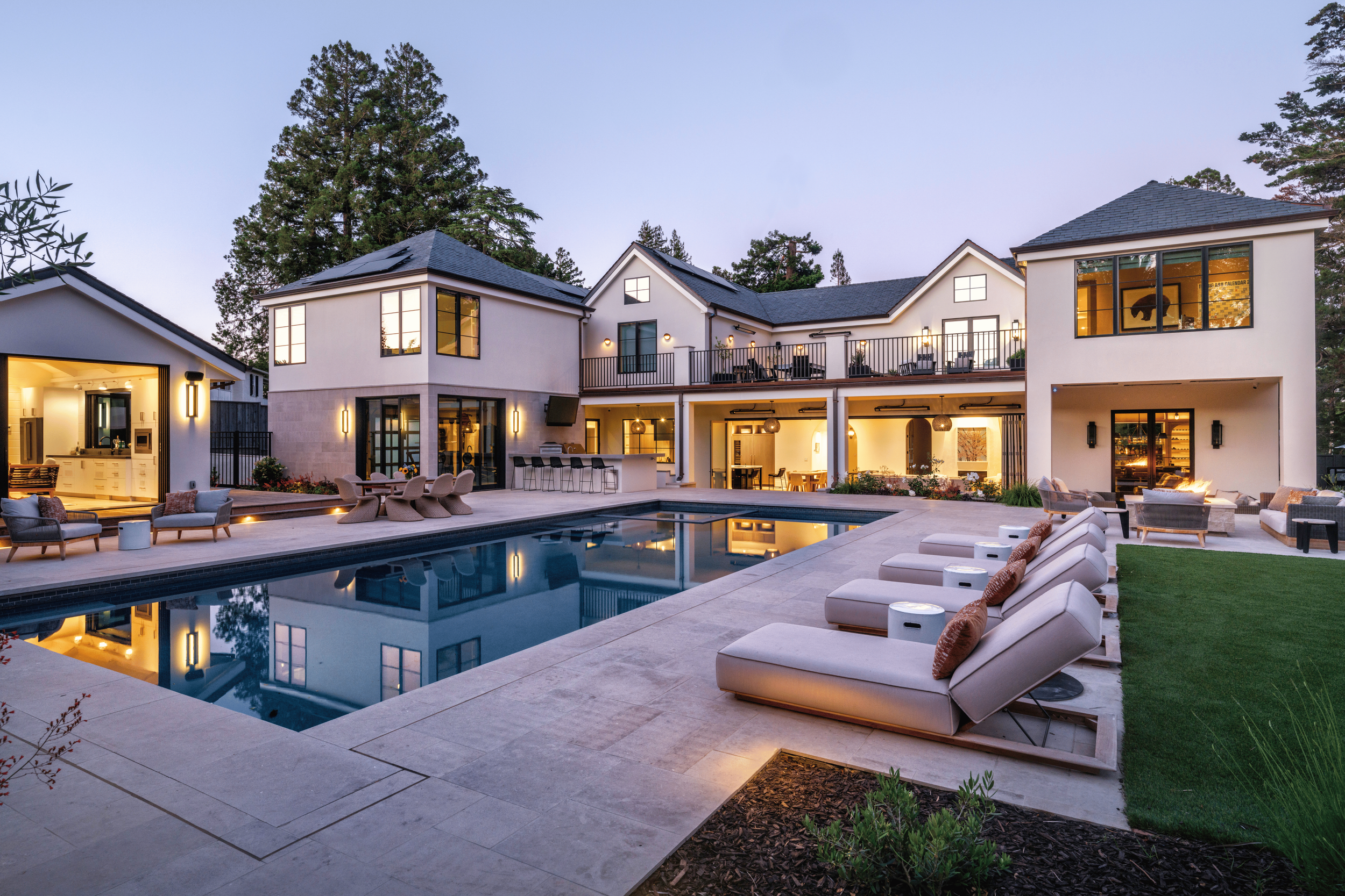 Luxury modern house with a large swimming pool, outdoor lounge chairs, and patio furniture, illuminated at dusk.