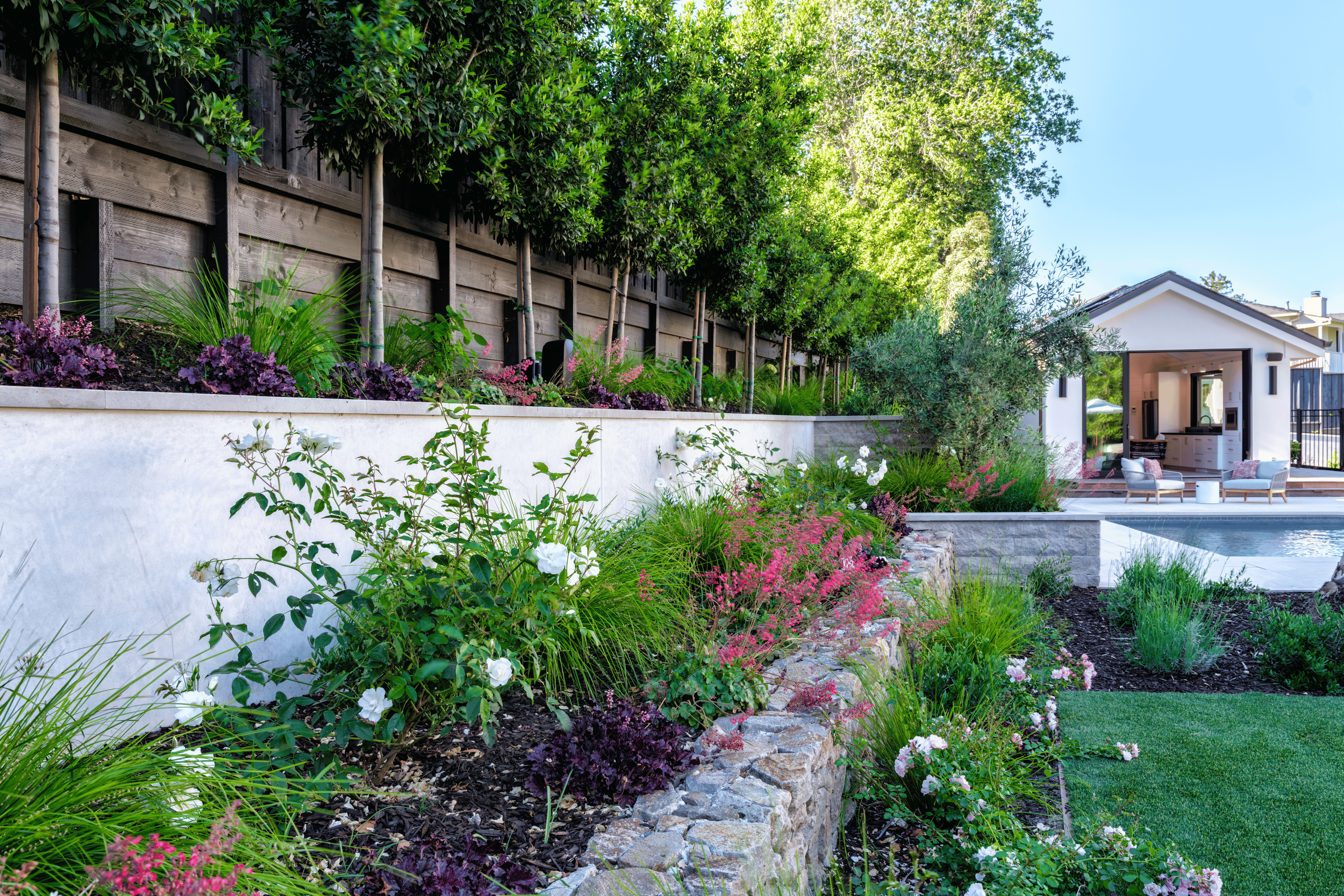 A landscaped backyard with a wooden fence and a variety of plants, trees, and flowers, adjacent to a modern outdoor living area with seating and a pool.
