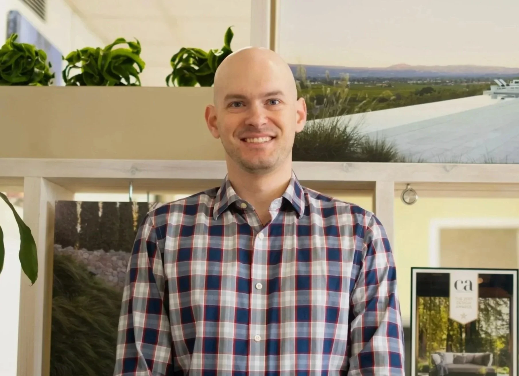 A man with a bald head and a checkered shirt smiling indoors with framed landscape photographs and green plants in the background.