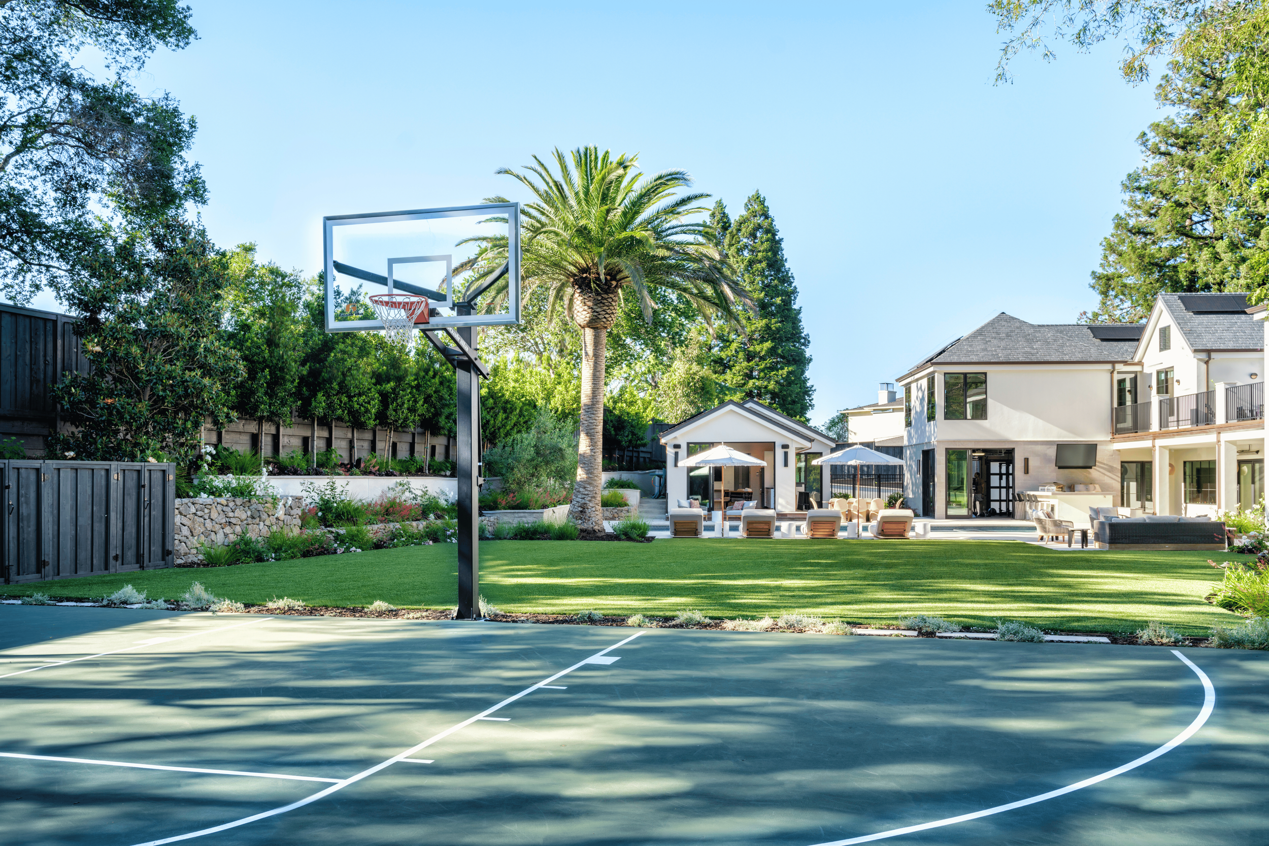 Residential backyard with a basketball court, green grass, palm trees, and outdoor seating area with umbrellas and modern house in the background.
