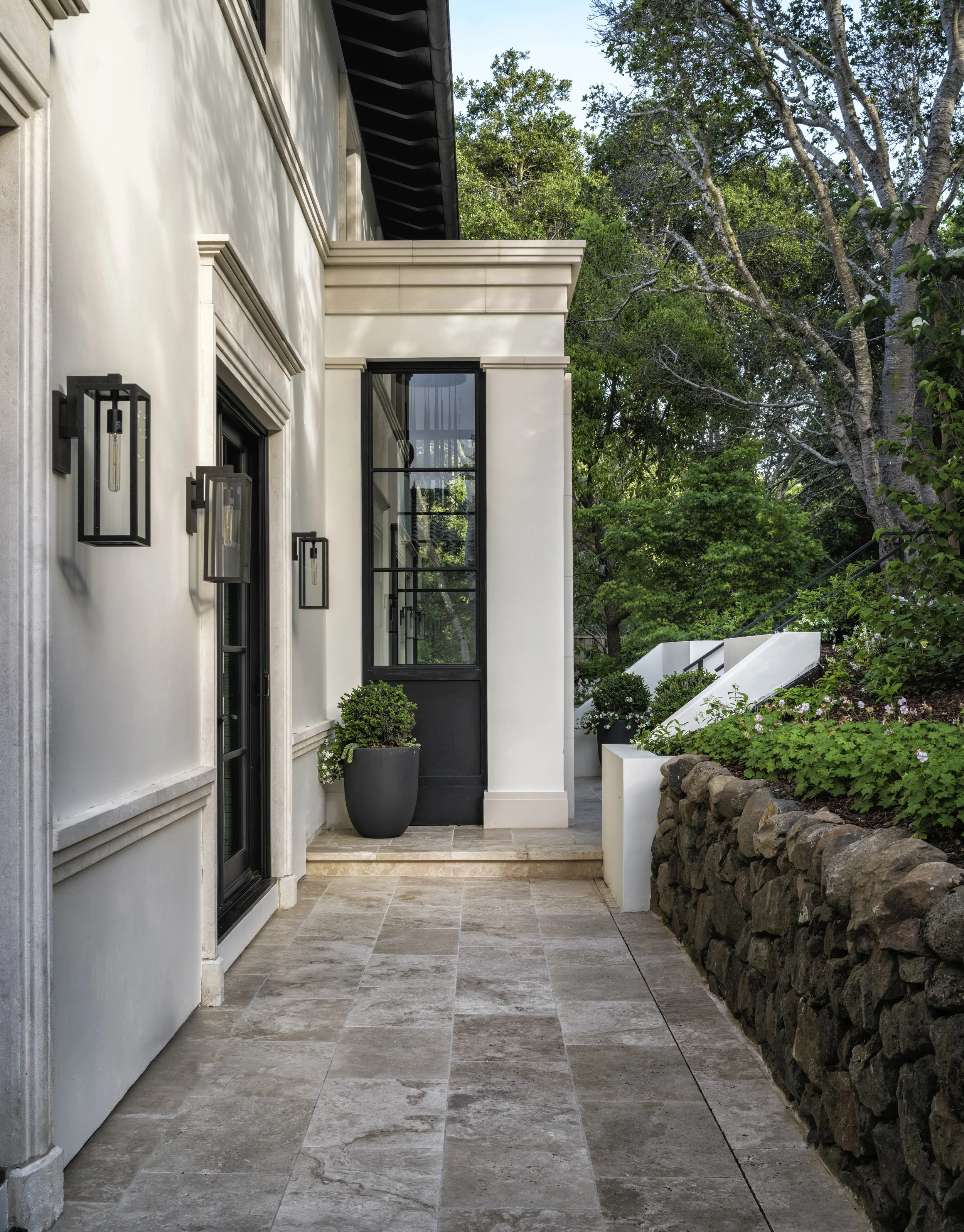 Exterior of a modern house with a tiled porch, black-framed glass windows, black wall-mounted lanterns, potted plants, a stone wall, and a set of white stairs leading into a lush green yard.