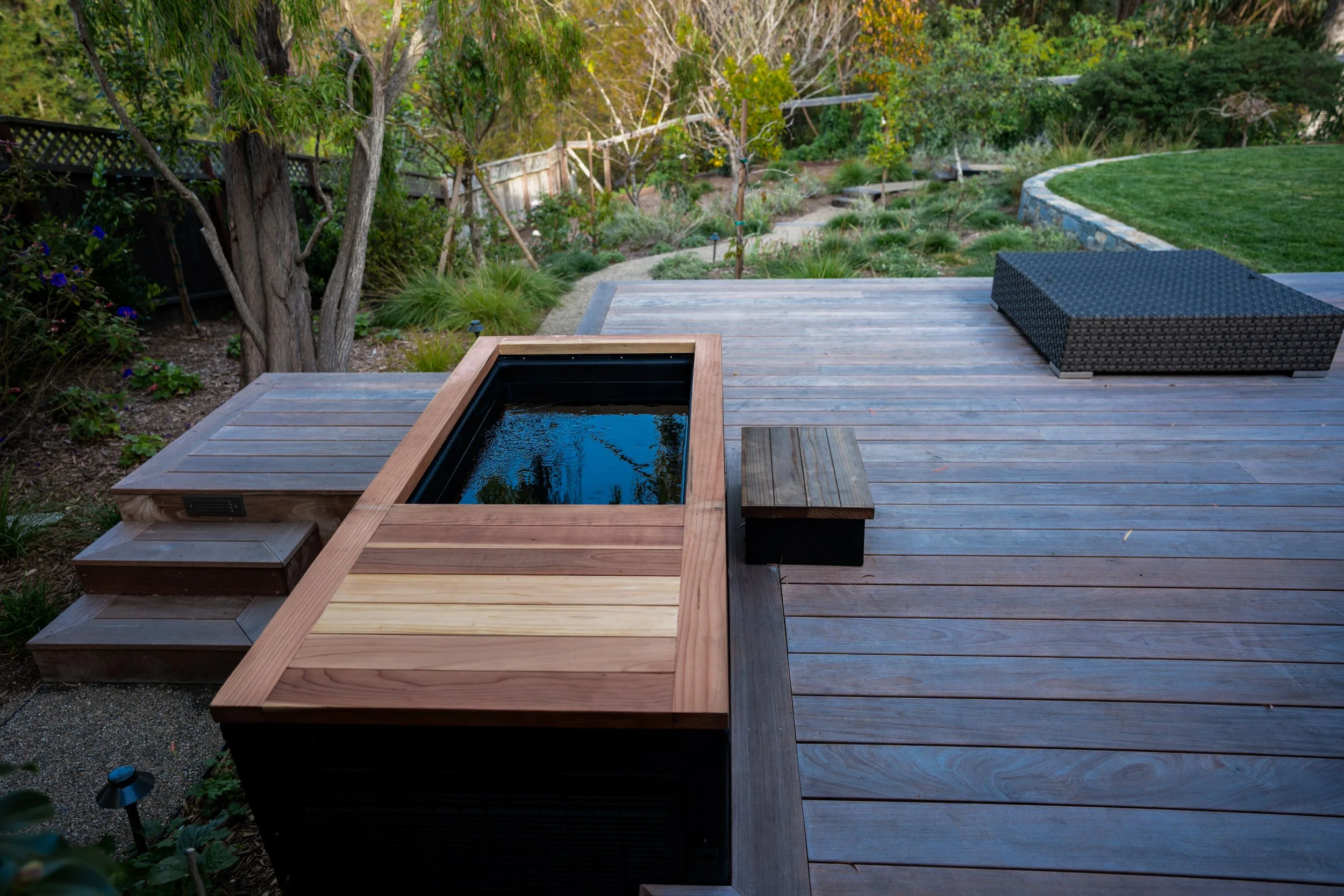 Outdoor wooden deck with a built-in black hot tub and a black wicker patio chair. The scene includes trees, shrubs, and a grassy lawn in the background.