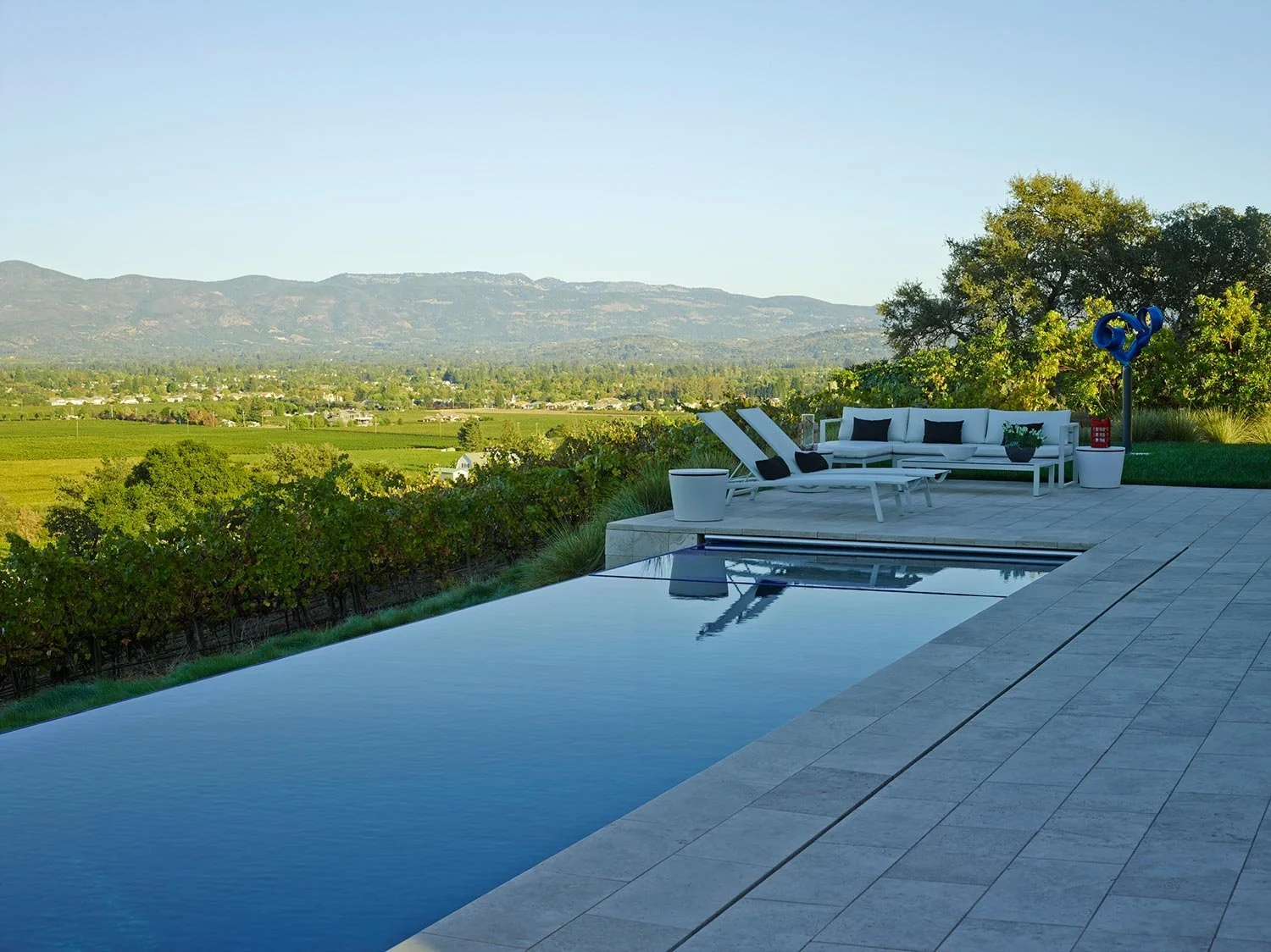 A backyard with a swimming pool, lounge chairs, a white outdoor sofa with black pillows, green plants, and a view of a landscape with fields and mountains in the distance.