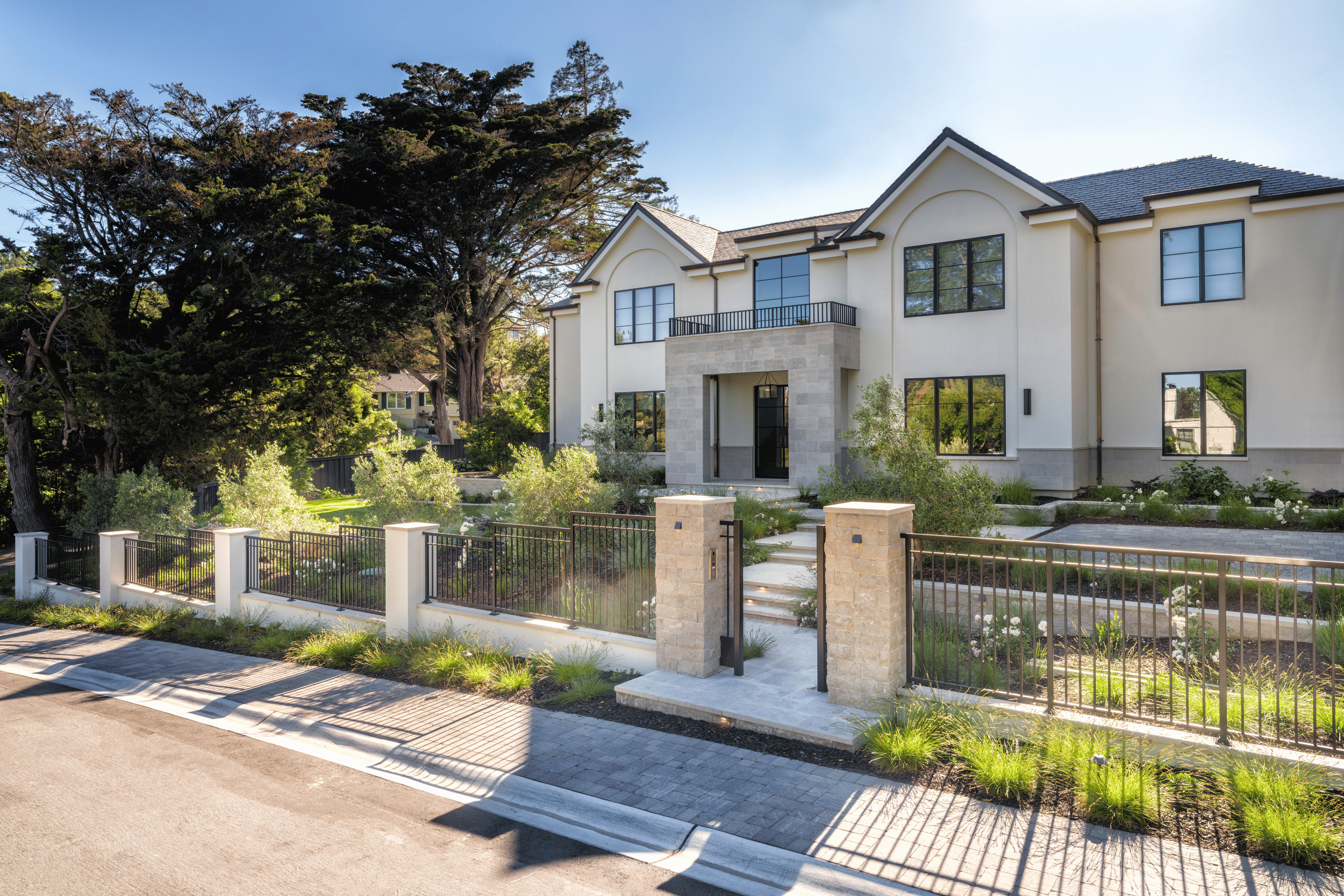 Modern two-story house with white exterior, large windows, and a gated front yard with landscaped greenery and trees.