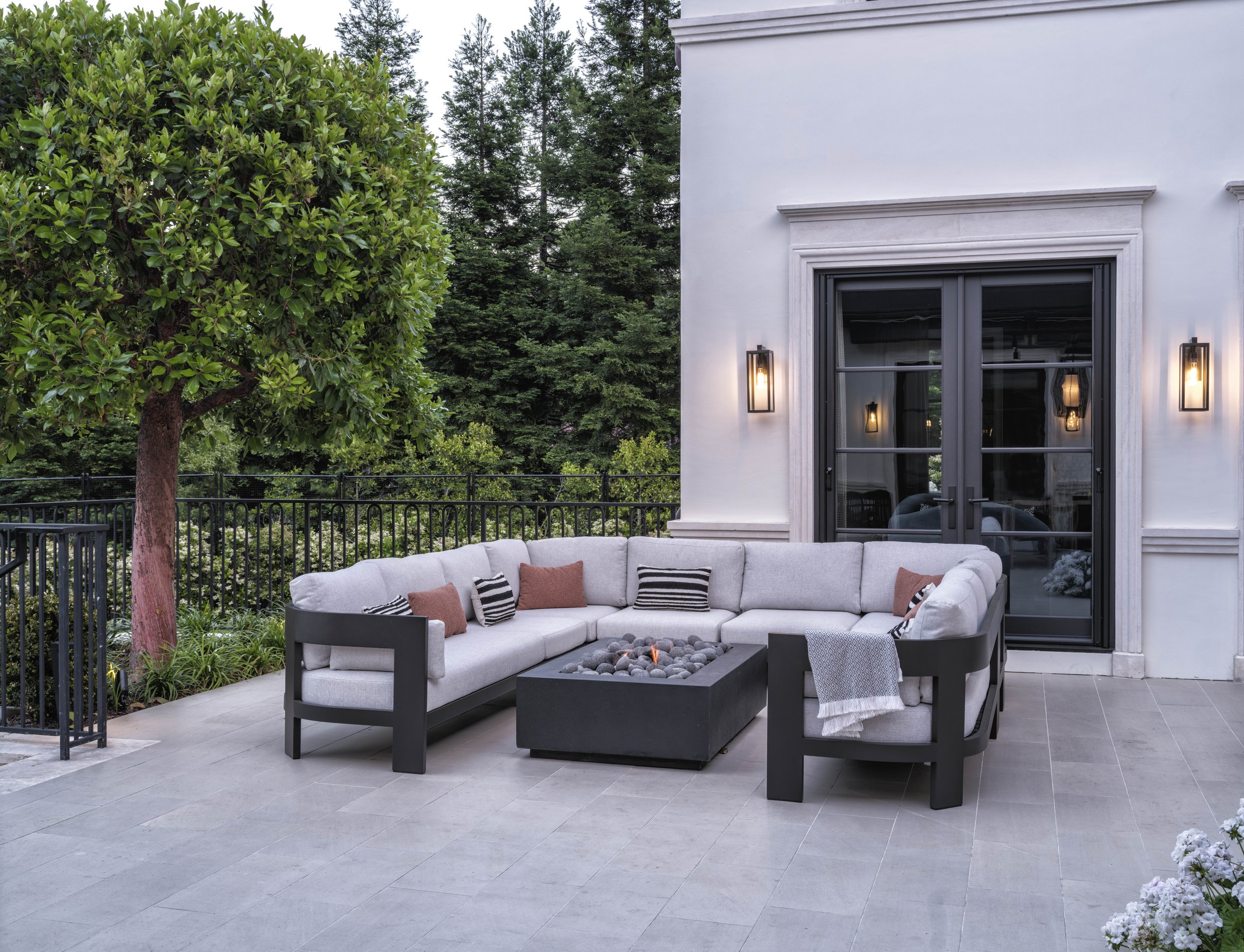 Outdoor patio with a large L-shaped sectional sofa with pillows, a fire pit table, trees, and a white house with black door and wall-mounted lights.