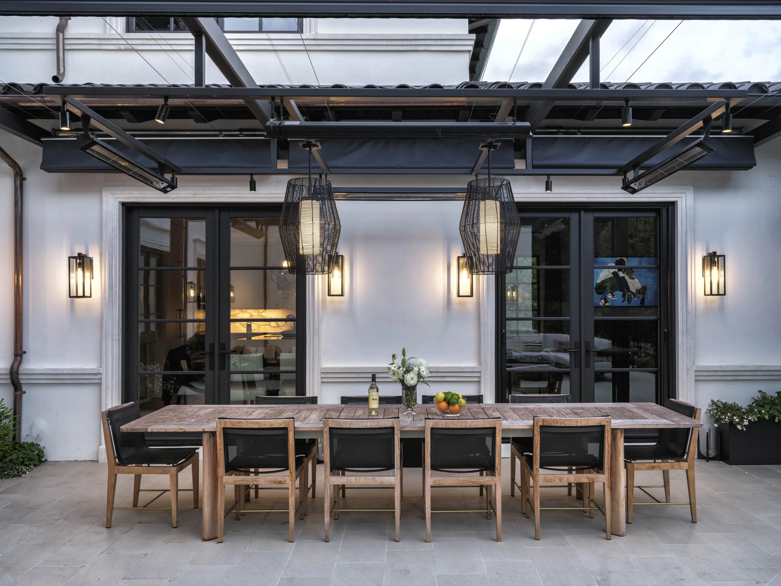 Outdoor patio with a large wooden dining table, eight black chairs, a vase with white flowers, a bottle of wine, and a bowl of oranges and limes. Two pendant lights hang above the table, mounted on a white wall with black-framed glass doors and moder