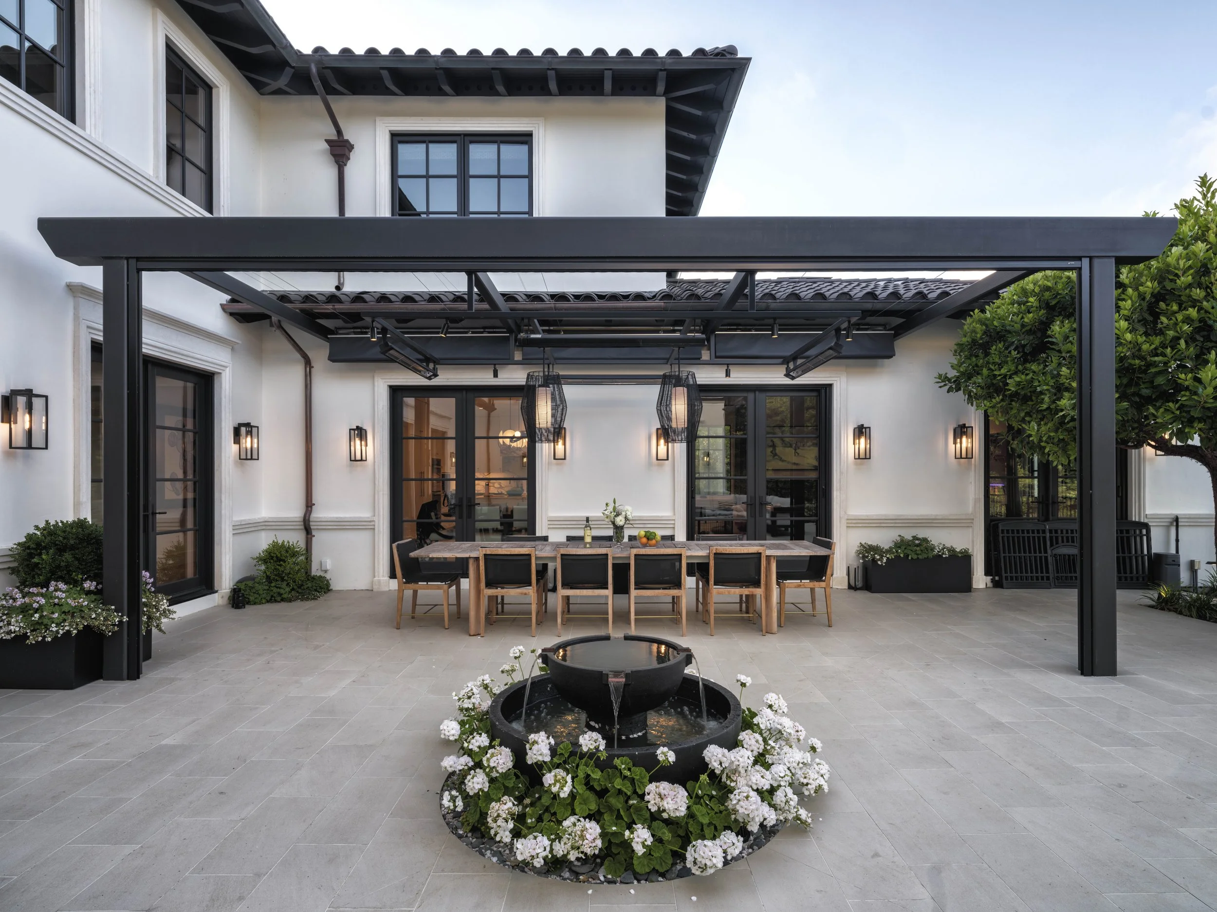 Backyard patio with a black pergola, a rectangular wooden dining table with eight chairs, a black flower fountain with white flowers, potted plants, and a white house with black framed windows and a glass door.