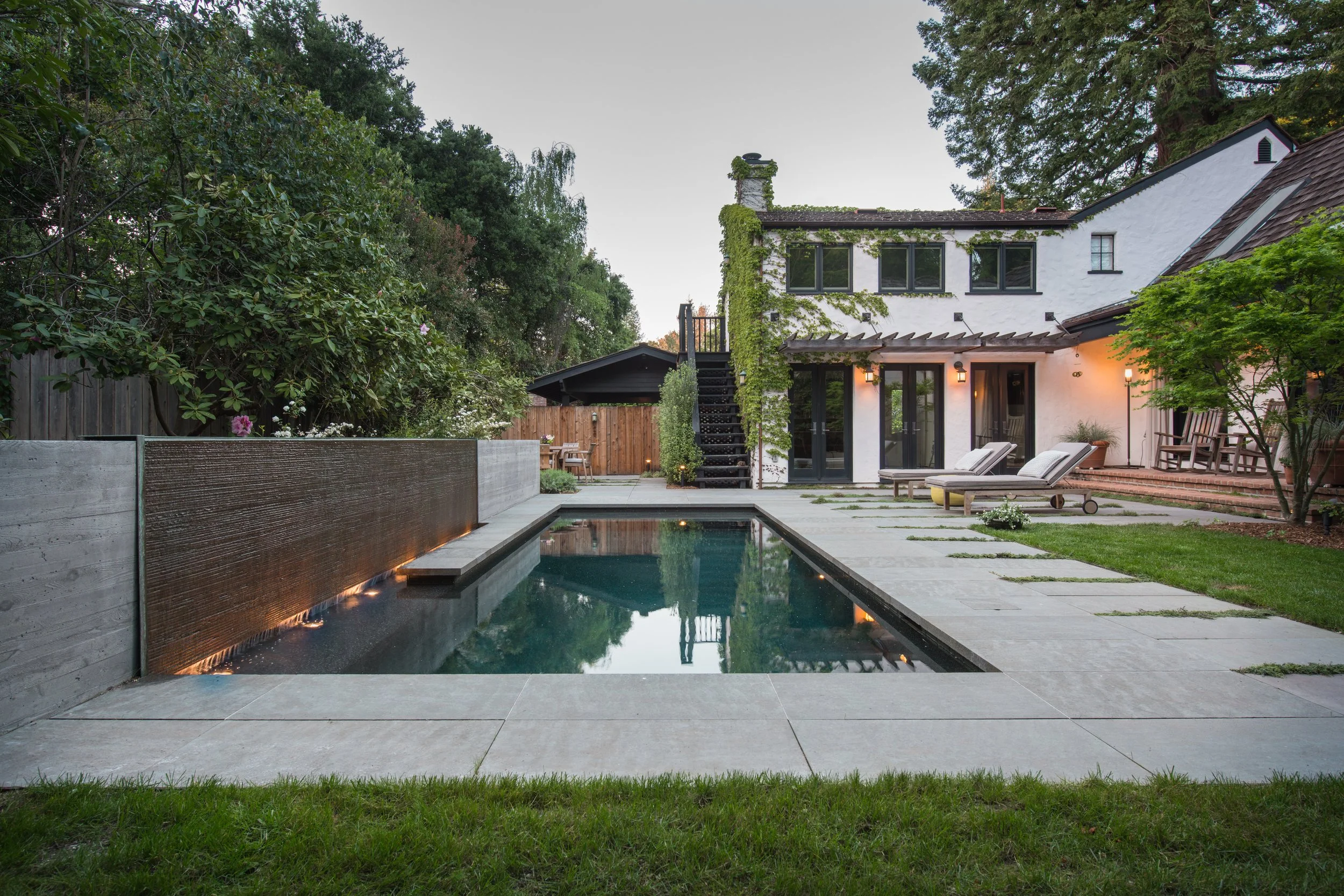 Backyard with a rectangular swimming pool, surrounded by a concrete patio, lounge chairs, and greenery. A white two-story house with black windows and a staircase is in the background.