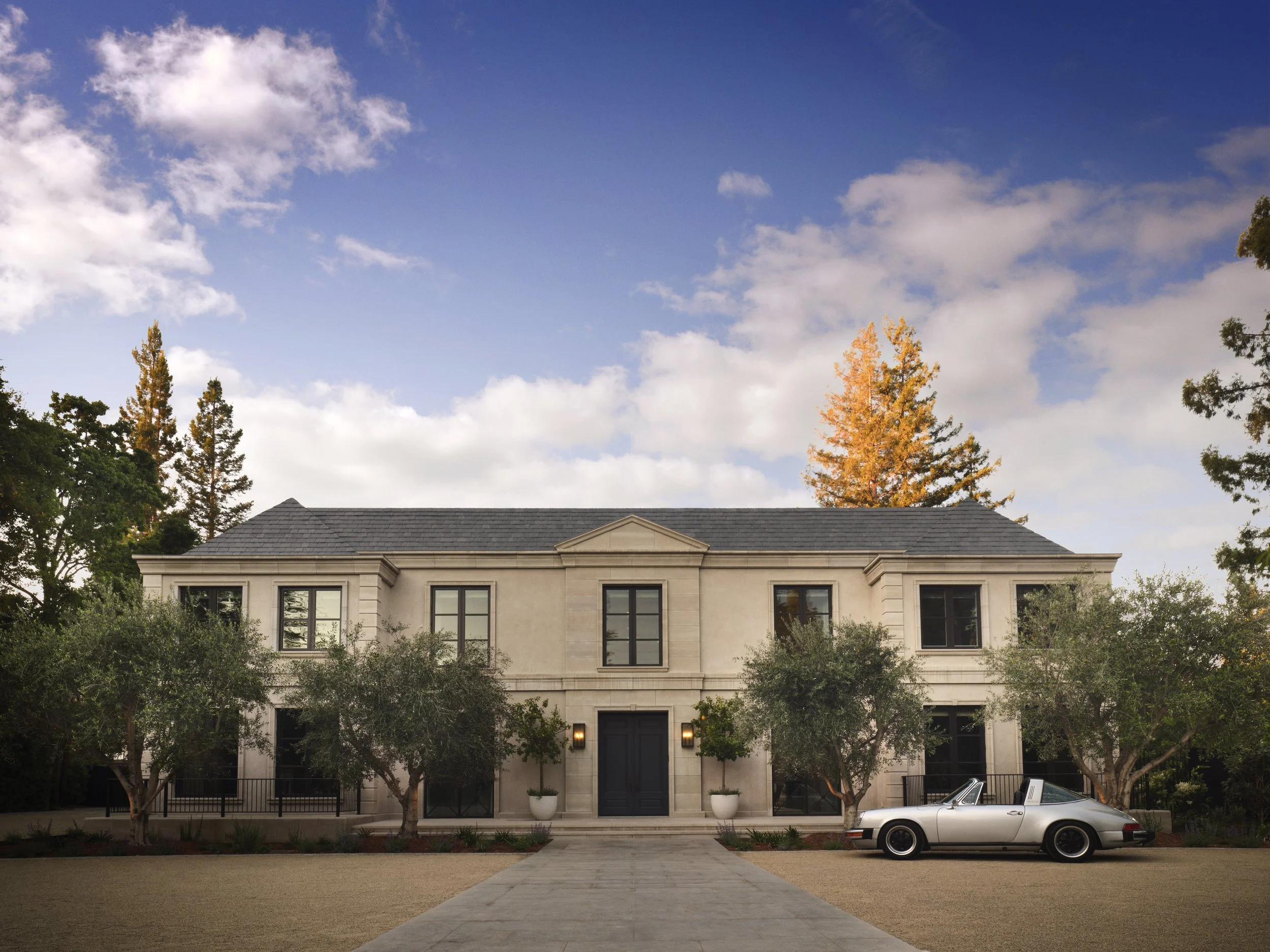 A large, elegant two-story house with a dark front door, surrounded by trees, with a vintage silver convertible car parked in the driveway, under a partly cloudy sky.