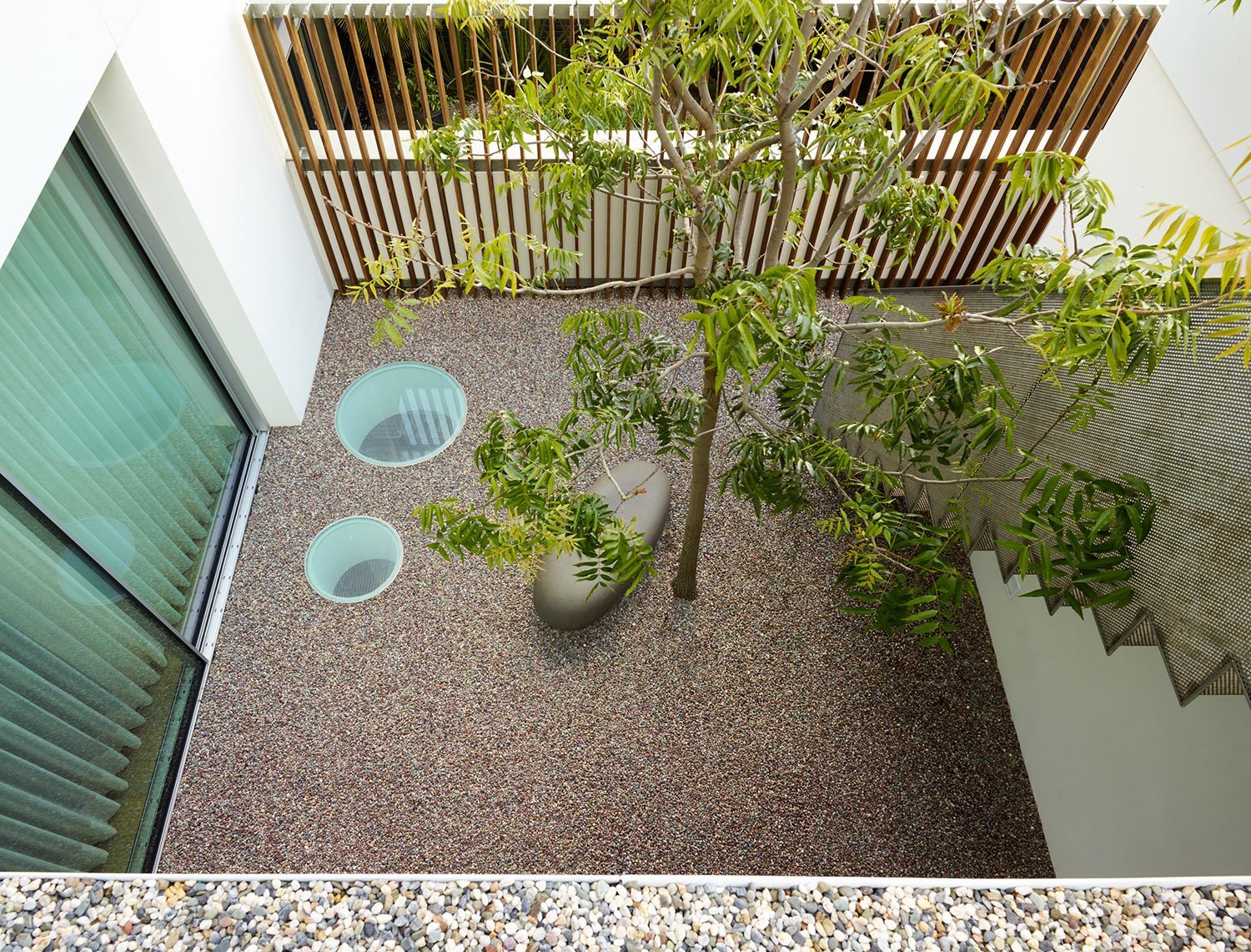 Top-down view of an outdoor balcony with two potted trees, two round floor vents, glass sliding door with green blinds, a wooden fence, and a staircase with a perforated metal railing.