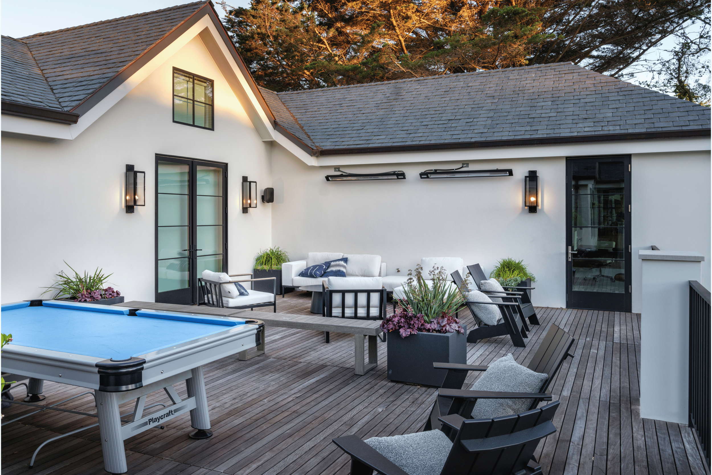 Outdoor rooftop patio with white and black furniture, potted plants, a foosball table, and a view of the house's white walls and sloped roof.