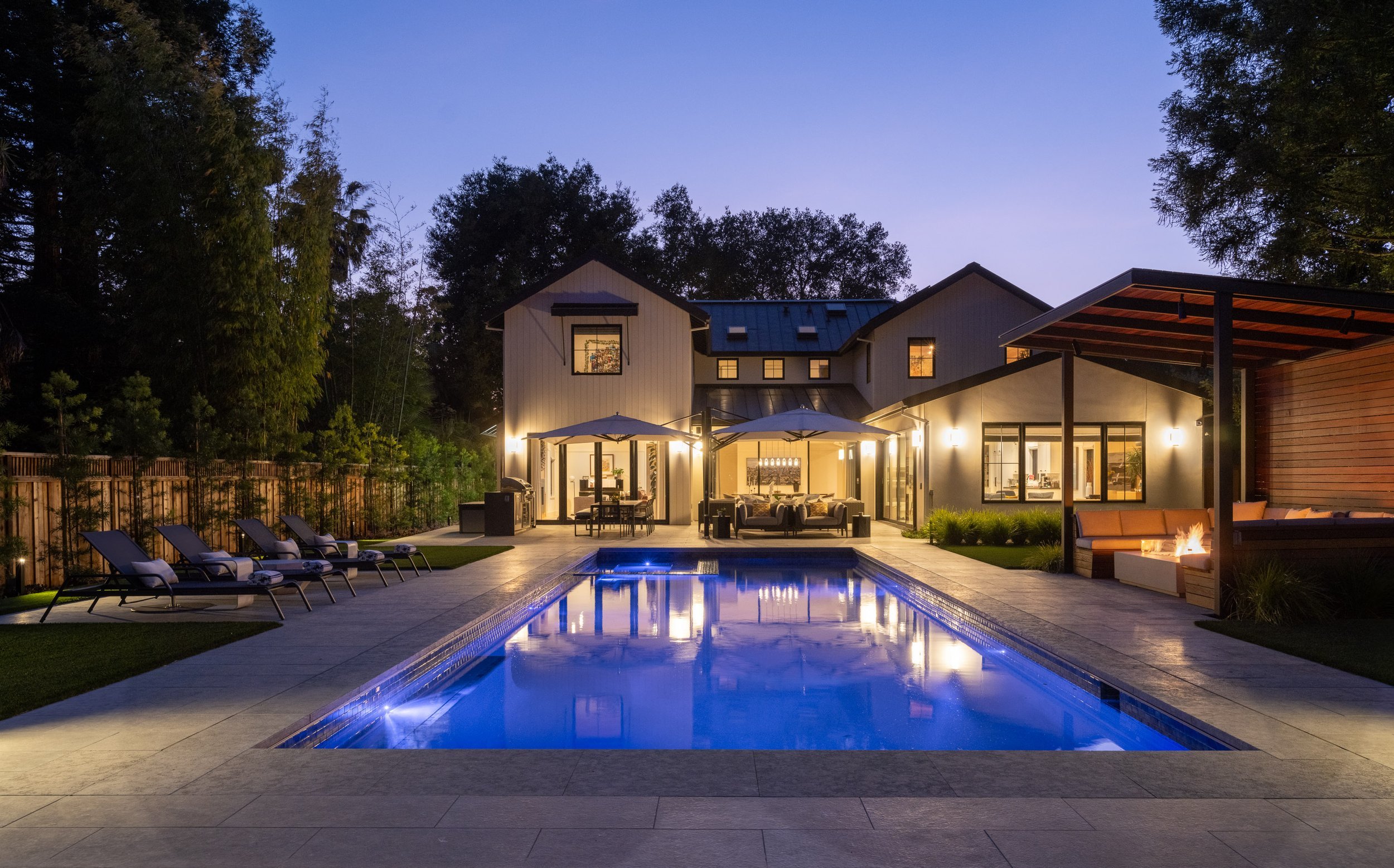 Backyard with a lit swimming pool, lounge chairs, outdoor seating area with umbrellas, and a modern house with large windows at dusk.