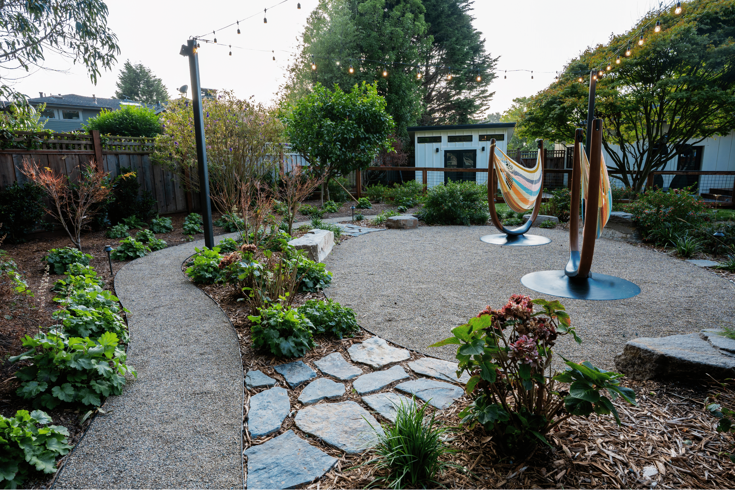 A backyard garden with a gravel area, a winding stone pathway, and a variety of plants and bushes. There are two hammock chairs with multicolored striped fabric hanging, and string lights overhead. A small shed is visible in the background, enclosed 