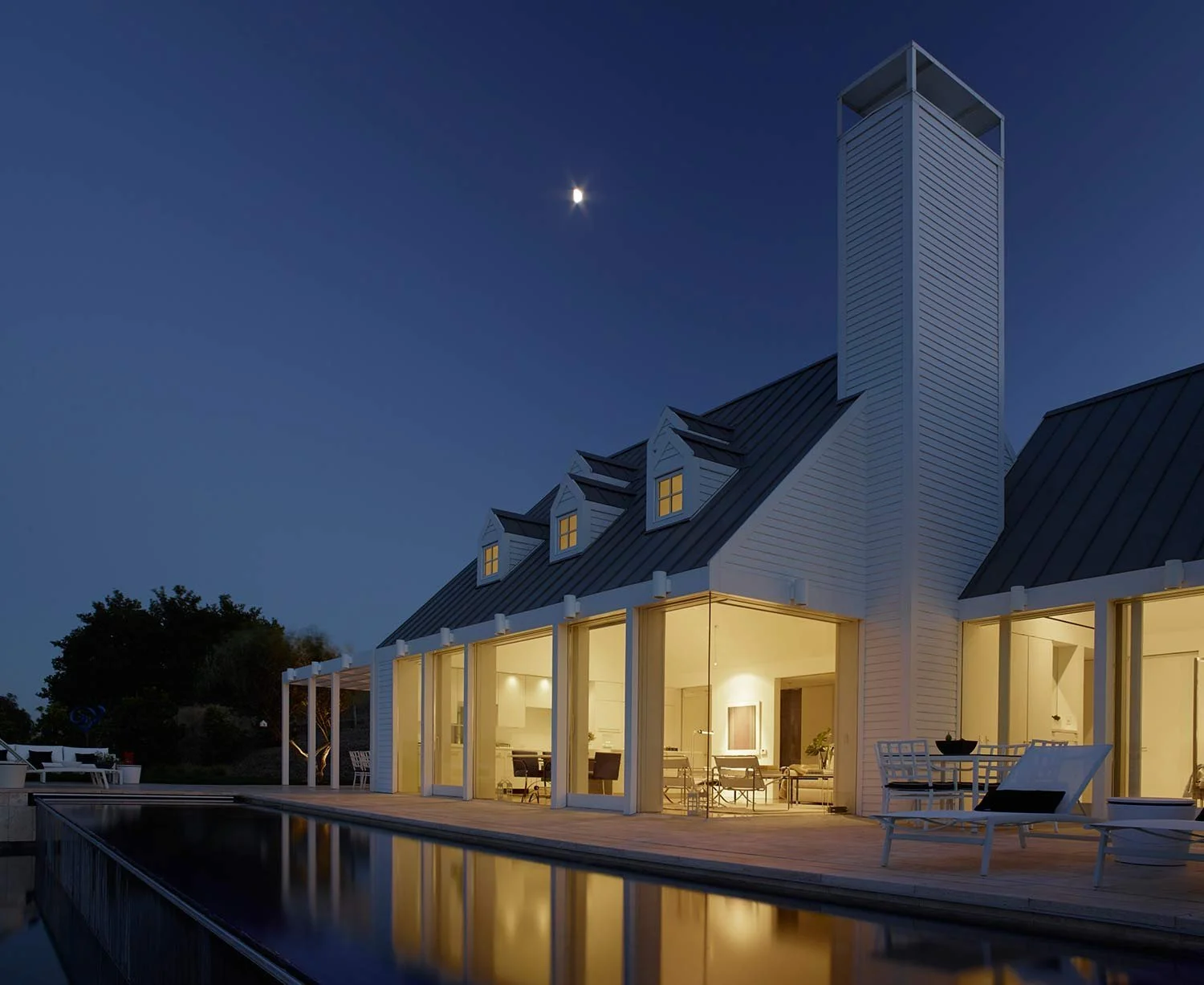 A modern house with a metal roof and large glass doors illuminated from within, sitting beside a reflecting pool at night. The sky above is clear, showing a bright moon.