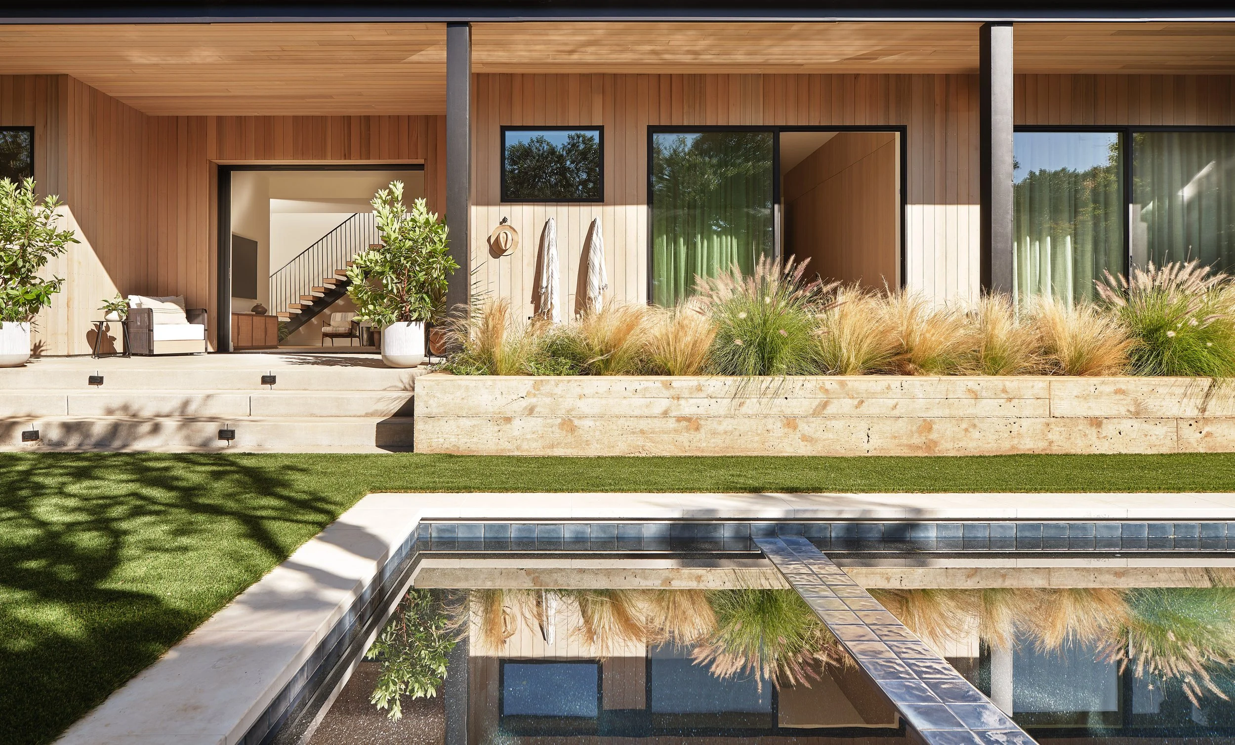 Modern house with a wooden exterior, large glass sliding doors, a small pool, and a landscaped yard with grass and ornamental grasses.