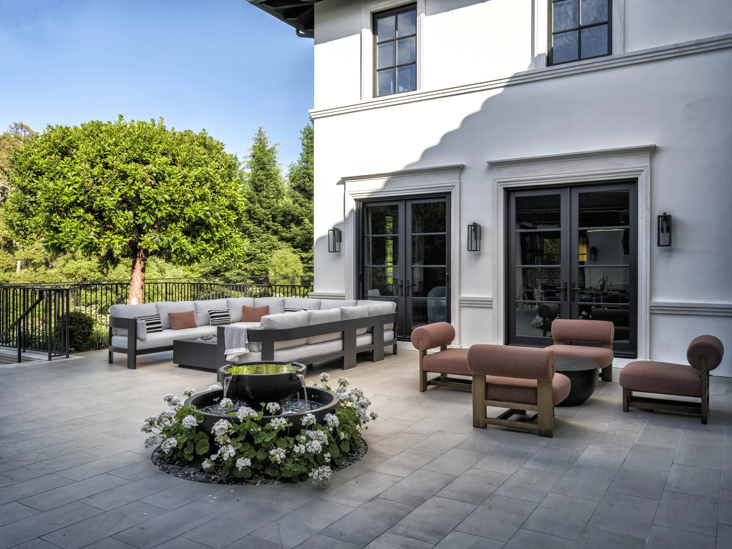 Outdoor patio area with a white sofa with cushions, pink and black chairs, a round black coffee table, a small fountain with flowers, and a large tree in the background. The area is enclosed by a black metal fence and is adjacent to the white exterio