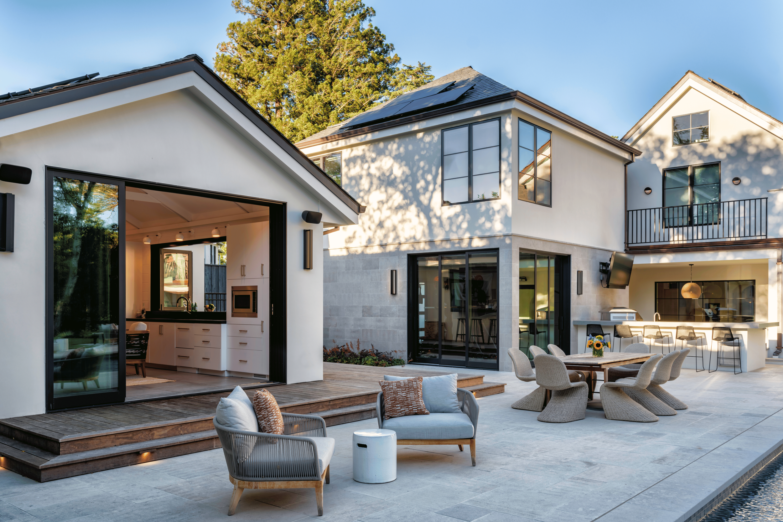Modern house with outdoor patio, lounge chairs, dining table, bar stools, kitchen area, and sliding glass doors, surrounded by trees.