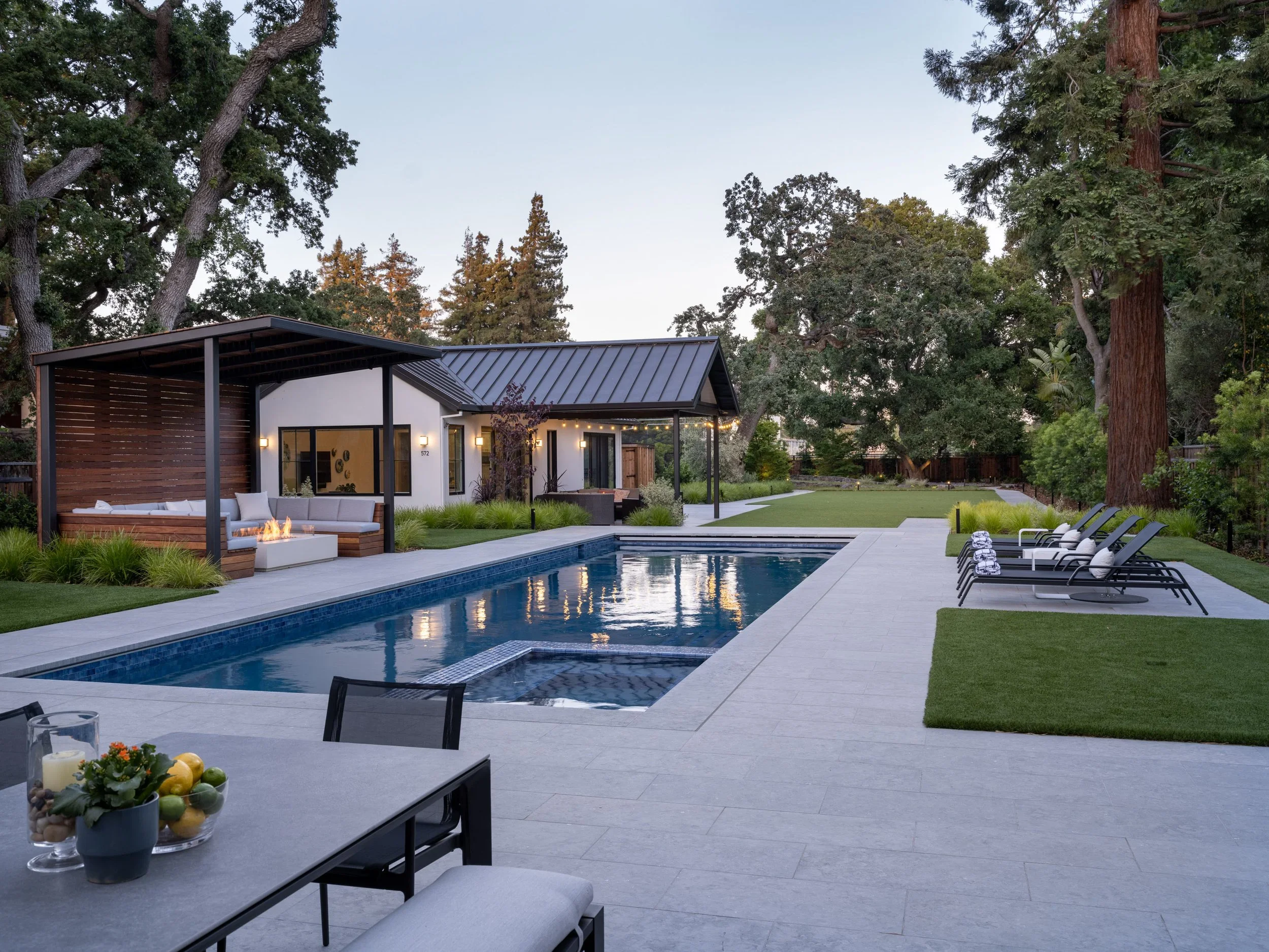 Backyard swimming pool with lounge chairs, a covered seating area with firepit, and surrounded by trees and greenery at dusk.