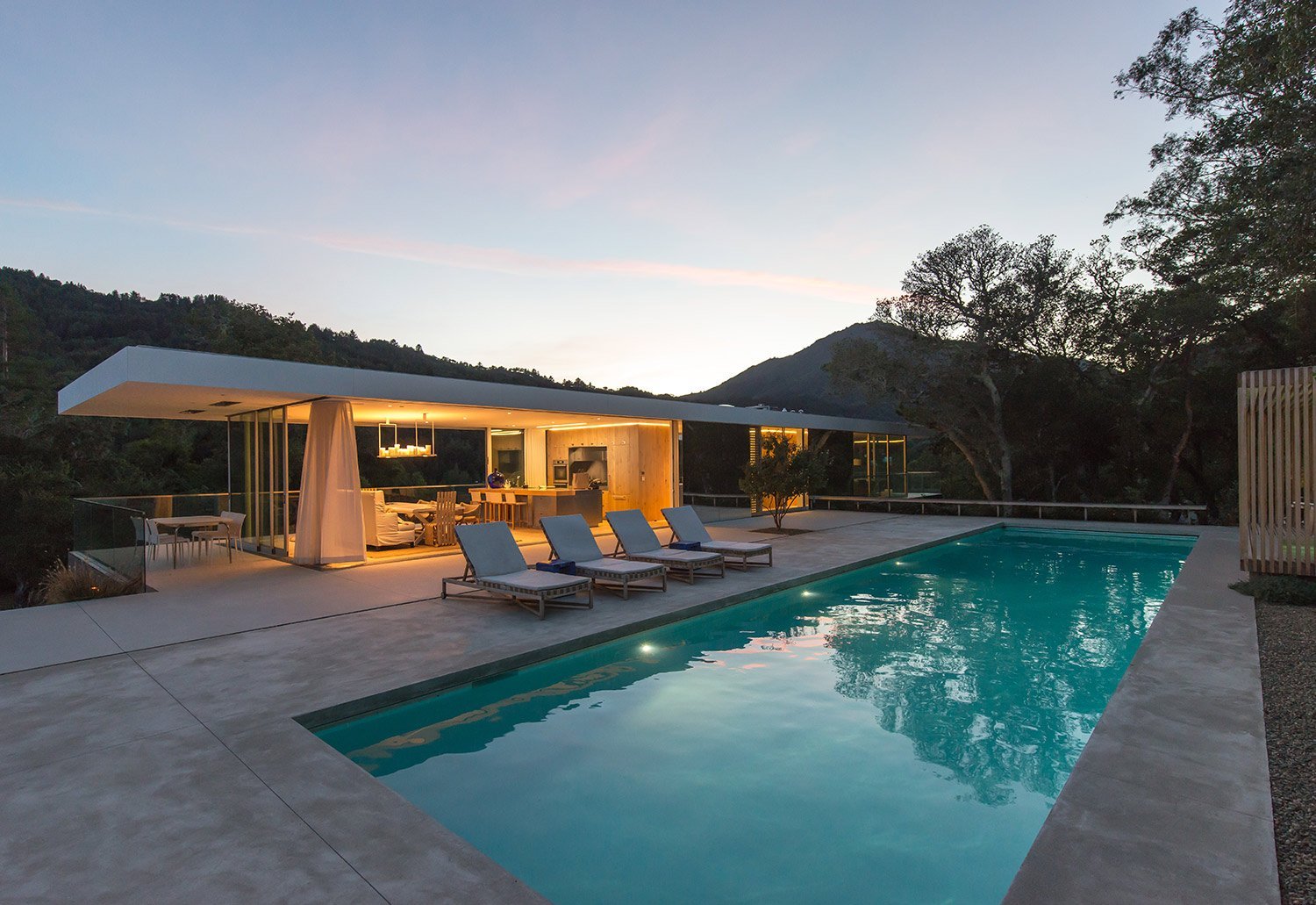 Modern house with large windows and an open living space, a swimming pool, four lounge chairs, and an outdoor dining area, set against a backdrop of trees and mountains at sunset.
