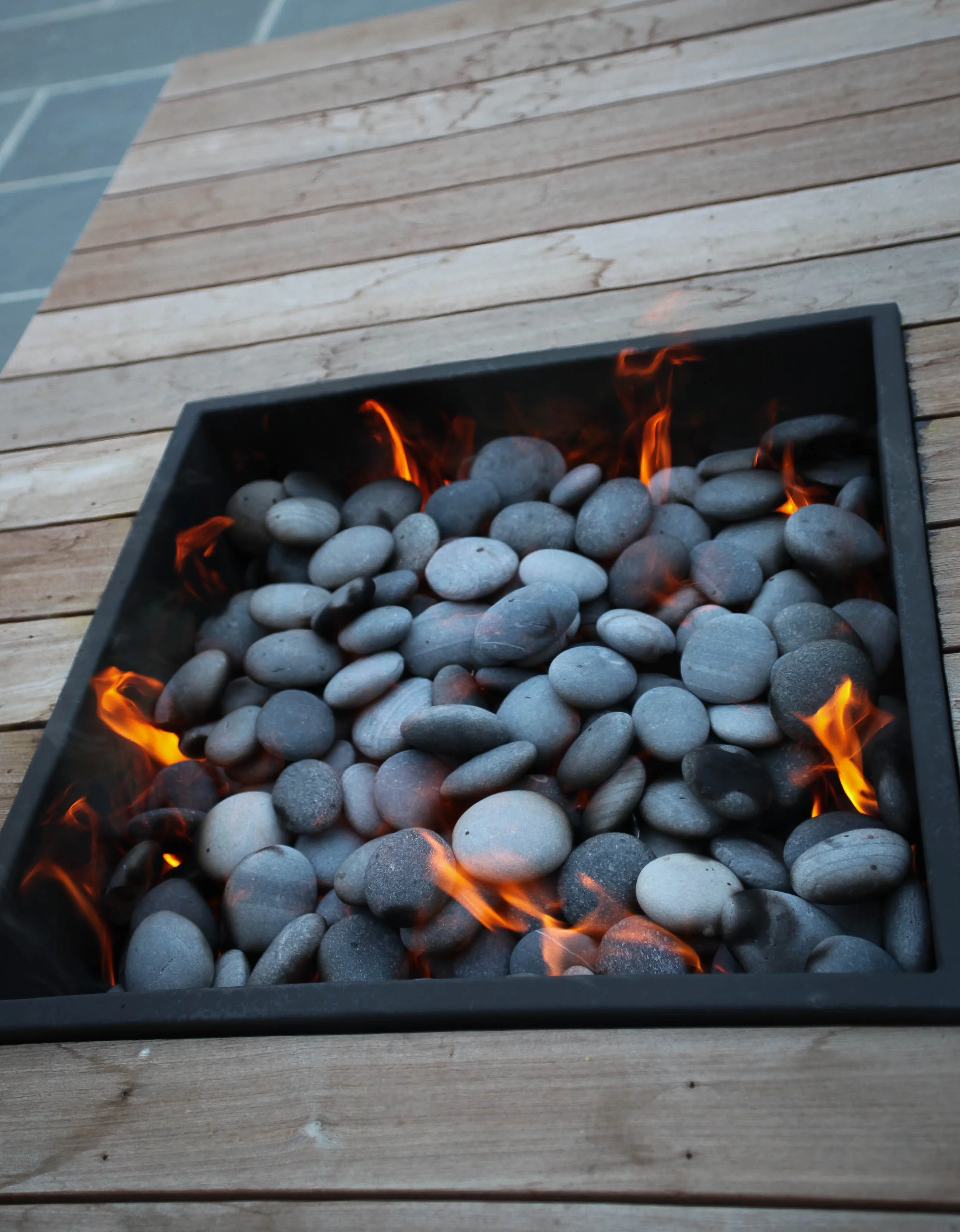 A fire pit filled with smooth gray stones, with flames rising among the stones, placed on a wooden deck.