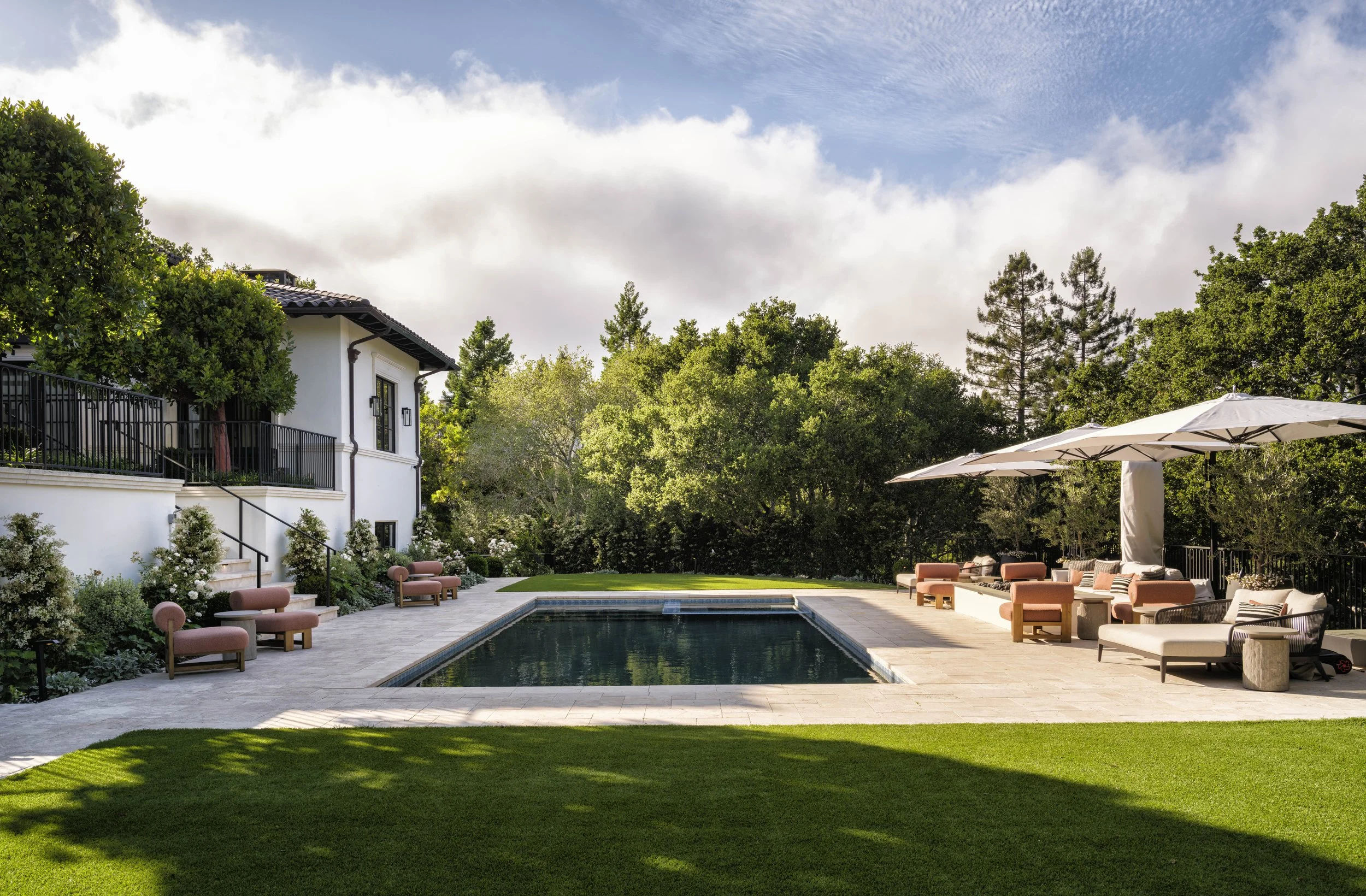 A backyard with a swimming pool, surrounded by lounge chairs, umbrellas, and lush green trees.