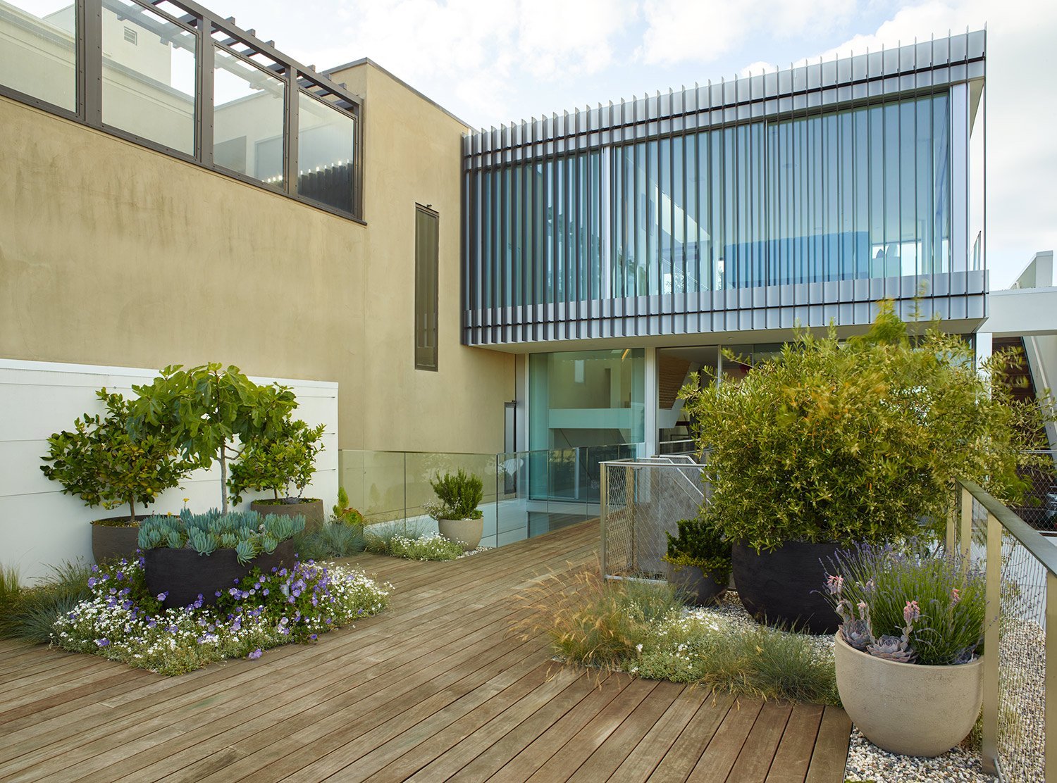 Modern house with a wooden deck and potted plants in the foreground, glass railings, and a balcony with vertical metal slats on a partly cloudy day.