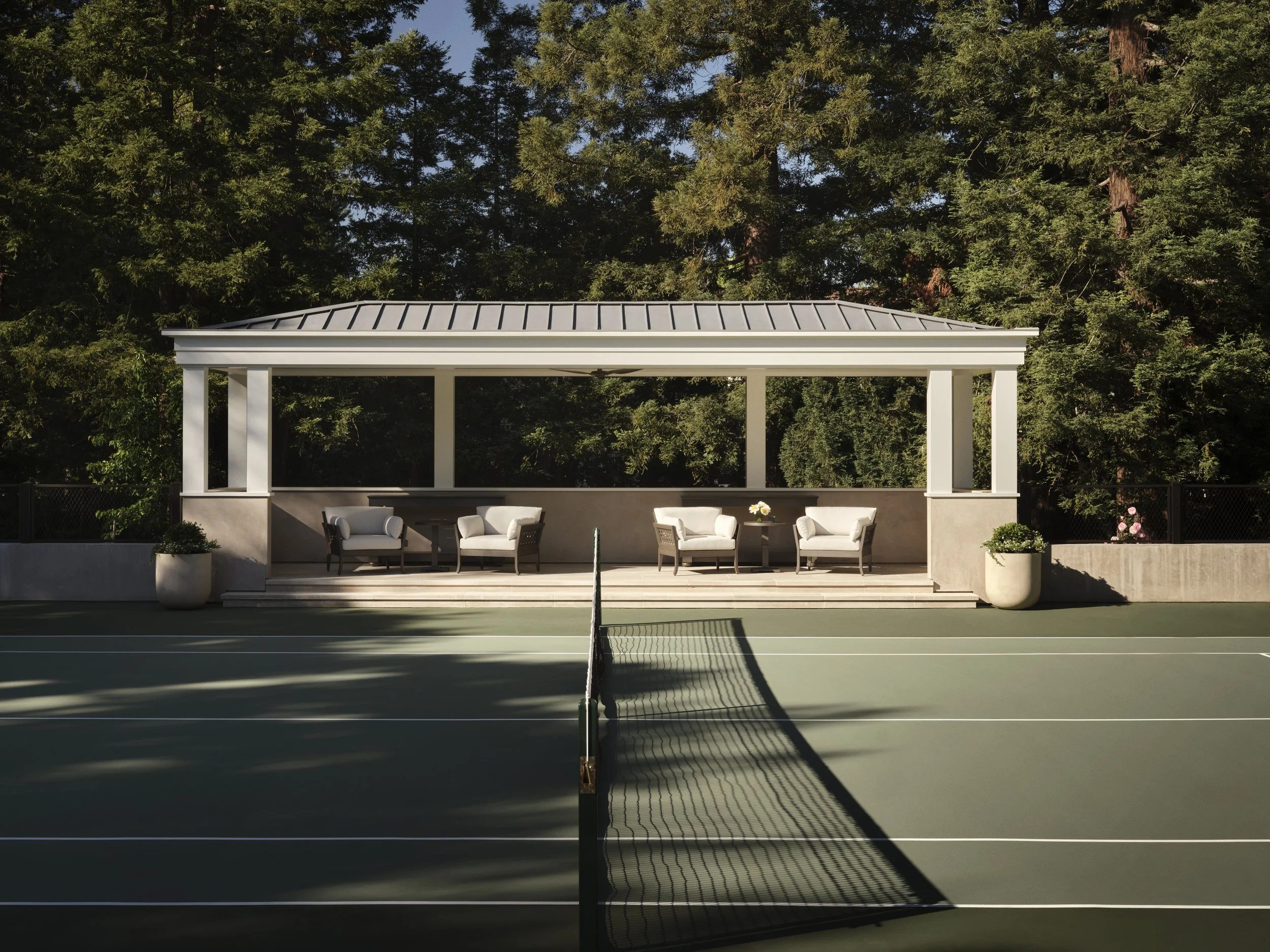 Tennis court with a pavilion and outdoor seating area, surrounded by trees.