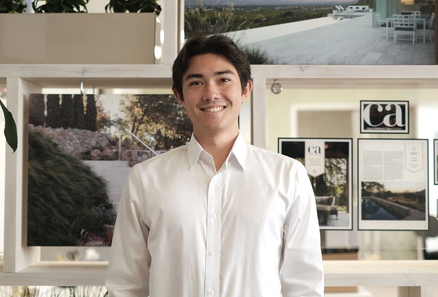 A young man with dark hair, wearing a white button-up shirt, standing indoors with a smile.