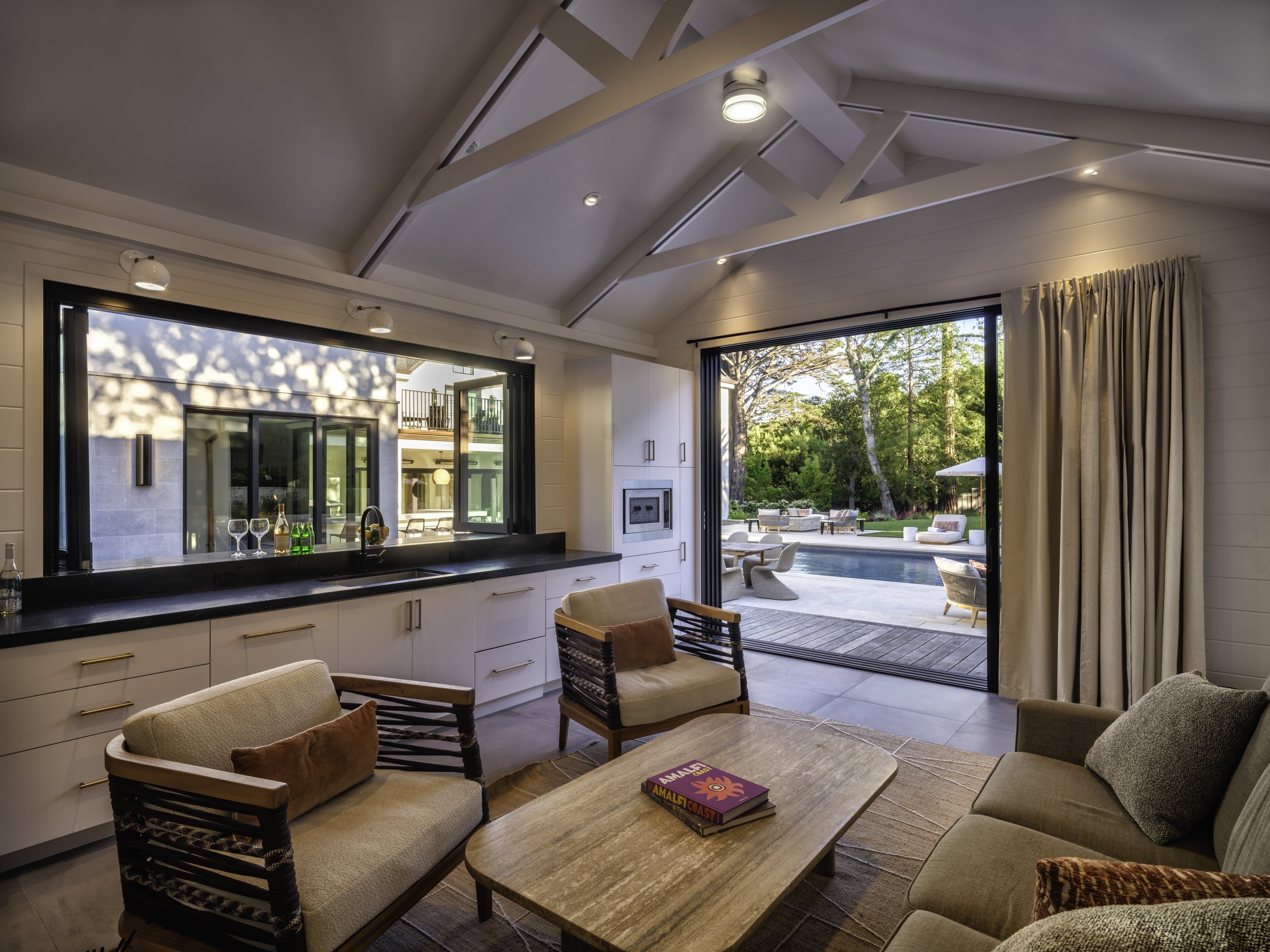 Living room with an open view of a backyard pool, outdoor seating, and trees, featuring a ceiling with exposed beams and a sliding glass door.