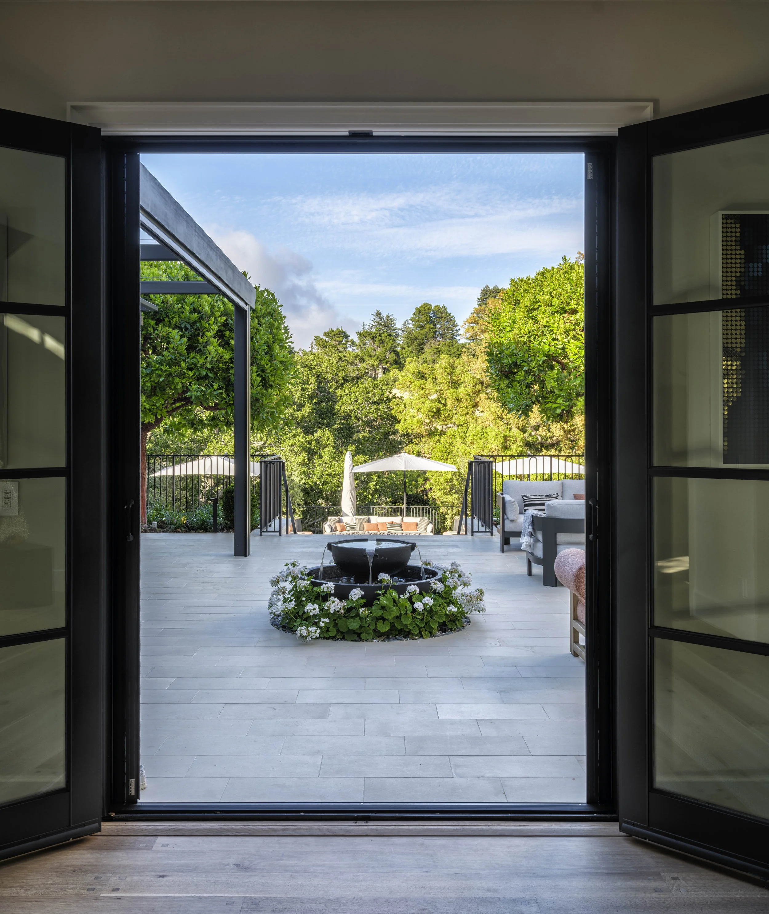 Open sliding glass door leading to a patio with outdoor furniture, umbrella, trees, and blue sky.