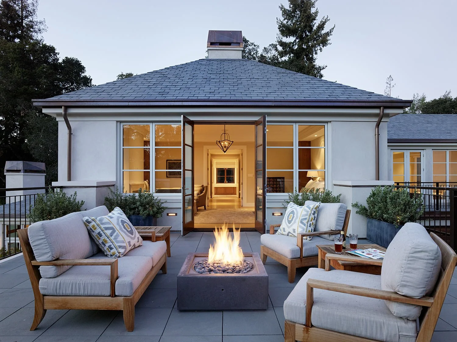 Luxury outdoor patio with fire pit, surrounded by sofas and armchairs, overlooking a modern house with large glass doors and interior lighting.
