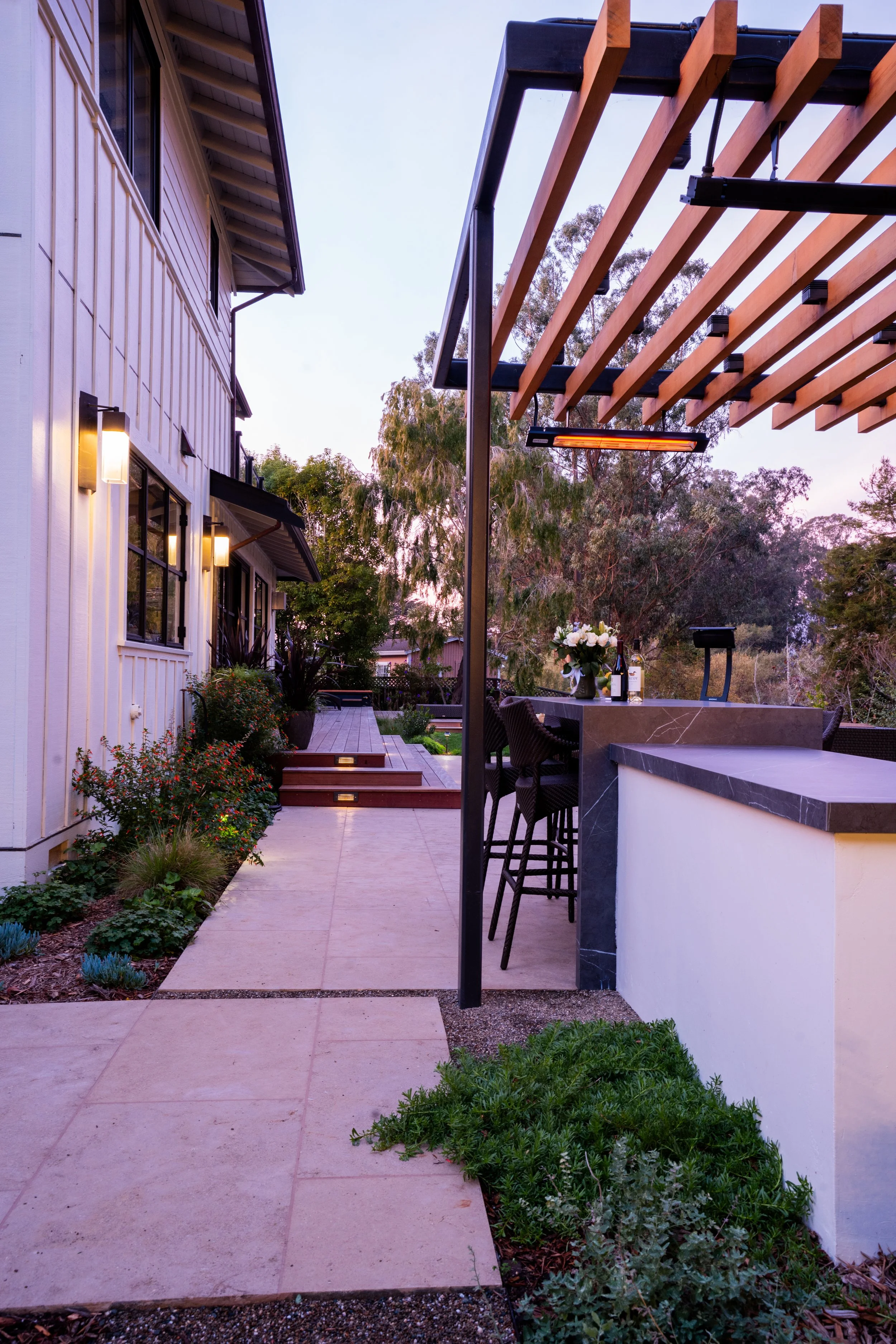 An outdoor patio area next to a house featuring a small bar with barstools, a flower arrangement, and a pergola overhead. The setting is during sunset with trees in the background.